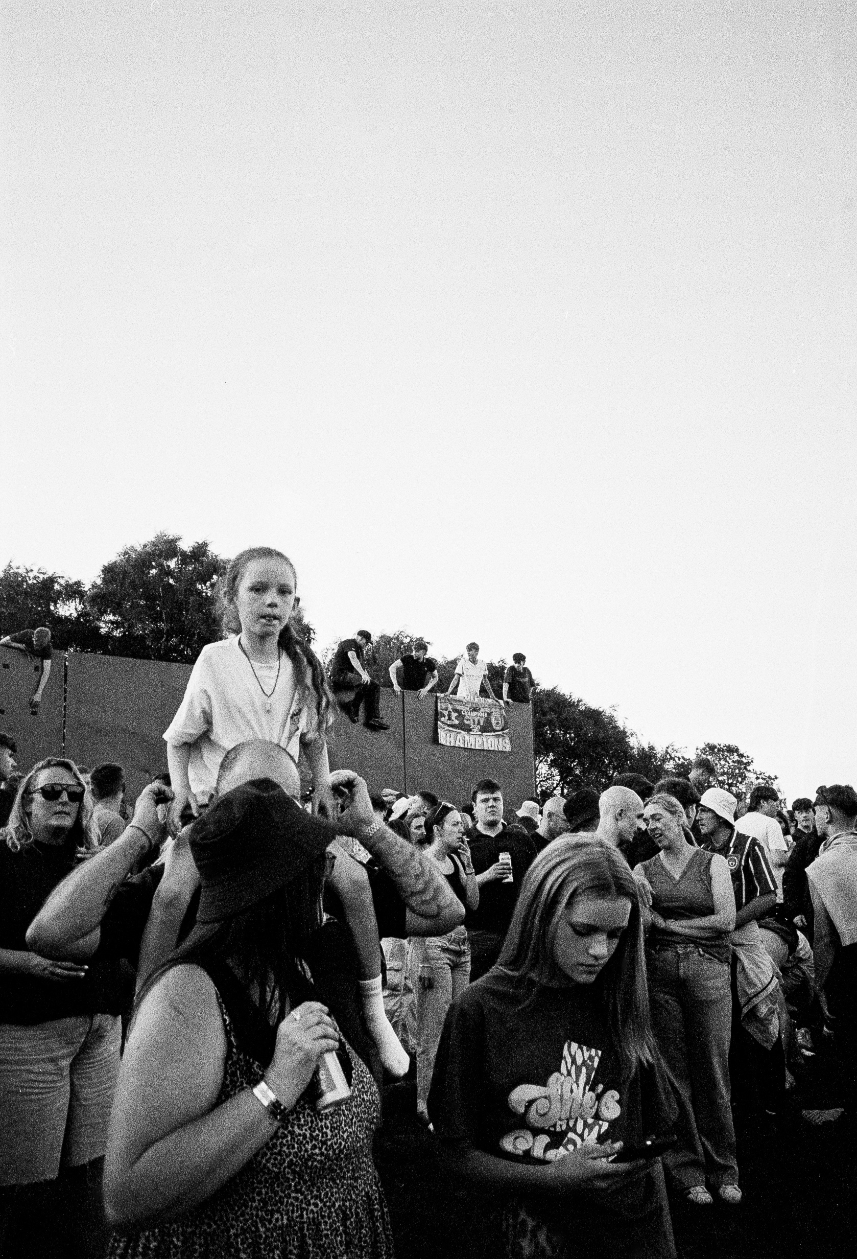 Child sits on adult's shoulders at crowded outdoor event, black and white image with trees and overcast sky in background.