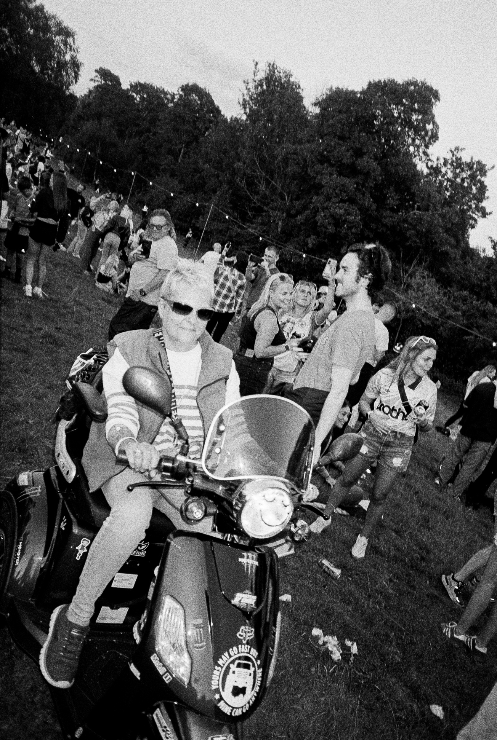 Black and white image of woman in sunglasses sitting on motorcycle at outdoor gathering with crowd and trees in background.