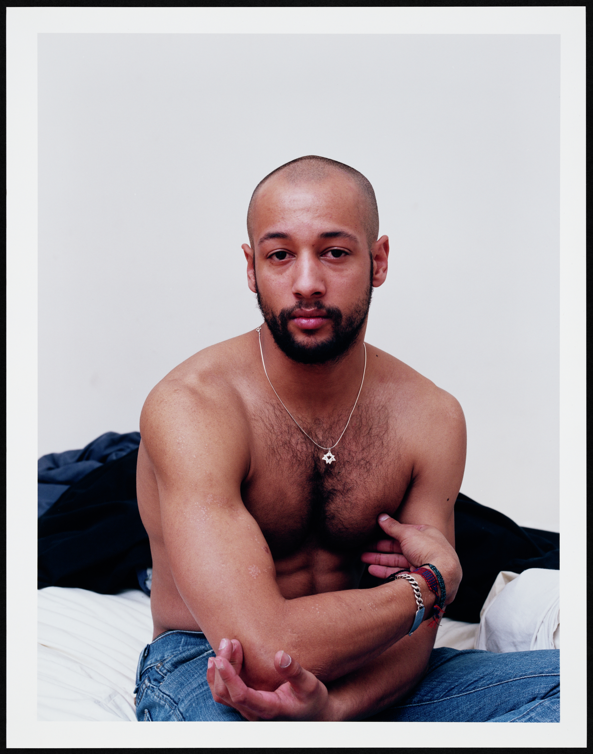 Shirtless man with a beard, sitting on a bed, arms crossed.