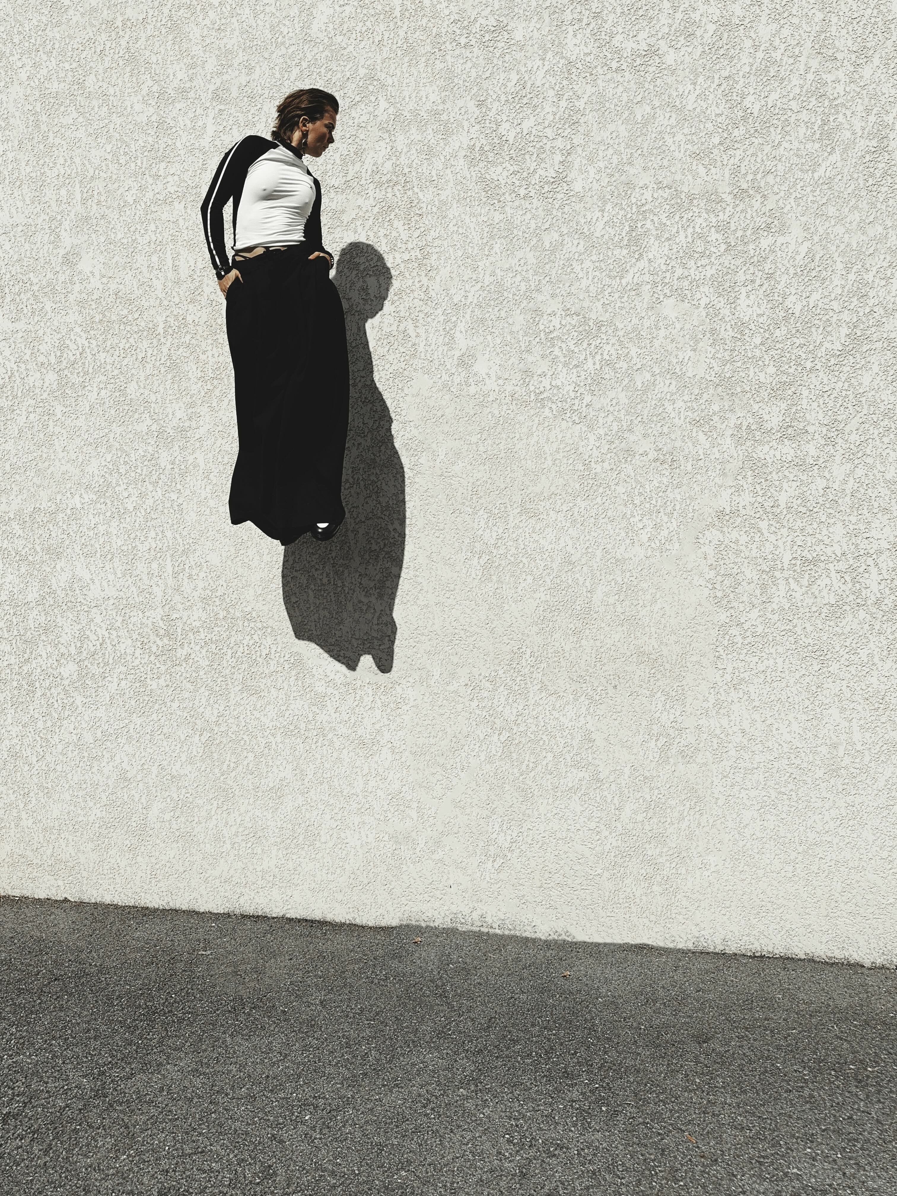 Man in white shirt and black trousers with braces stands on textured concrete surface, casting dark shadow on pale wall behind.