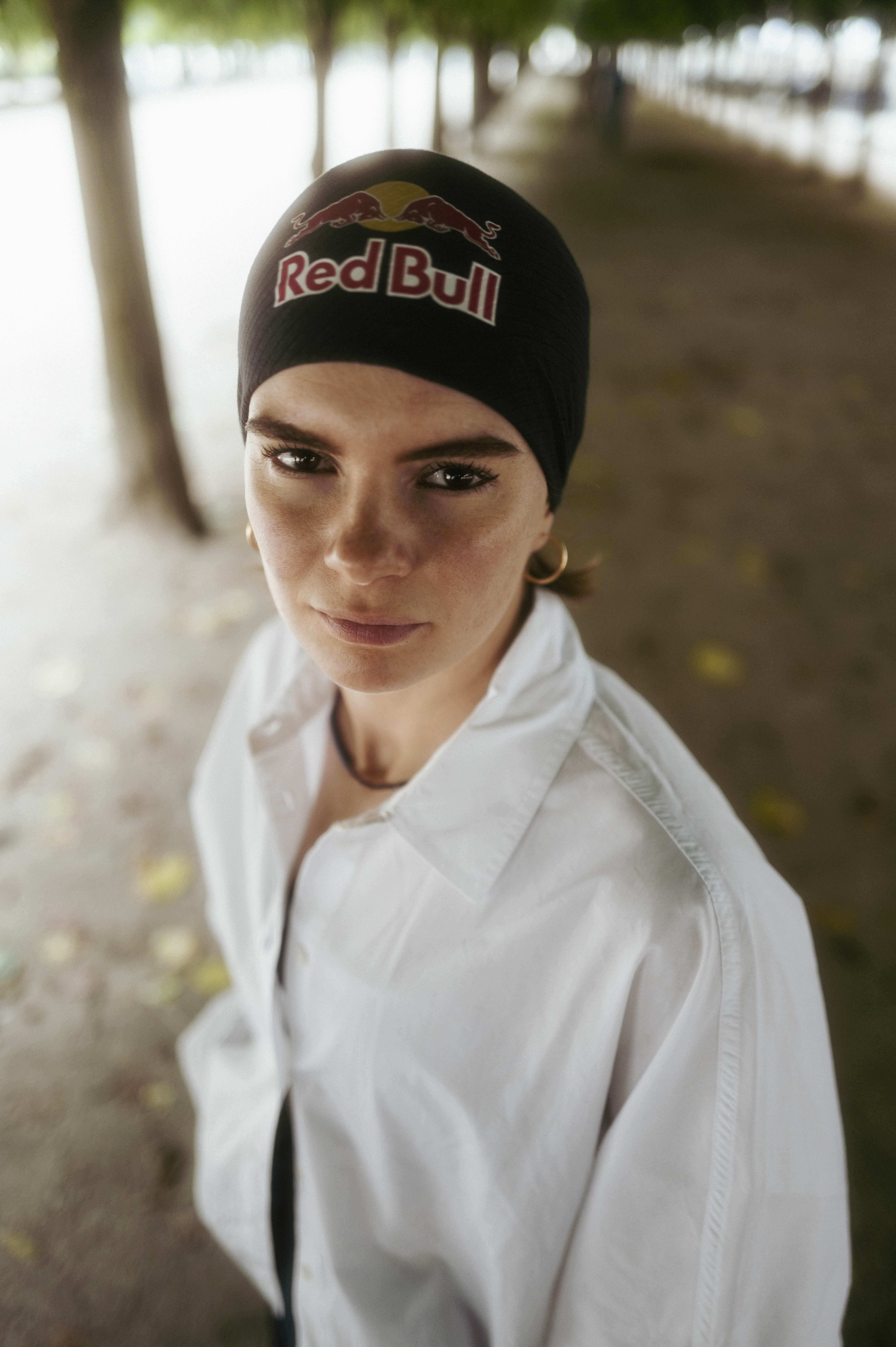 Woman wearing black Red Bull beanie and white zip-up hoodie, smiling at camera with blurred outdoor background of trees and pathway.