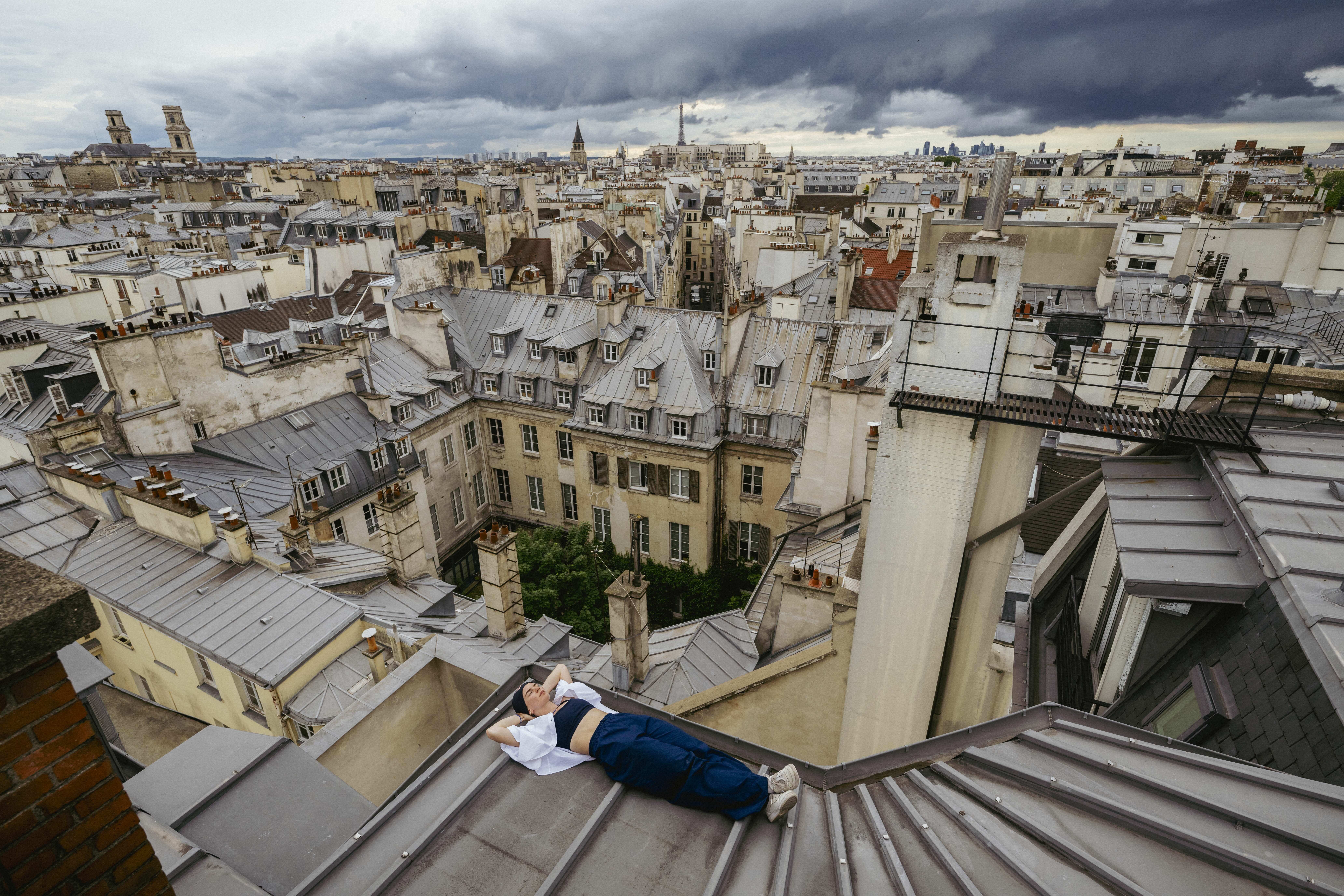 Person lying on grey metal rooftop with sprawling cityscape of cream and beige buildings below, dramatic grey and white clouds above.