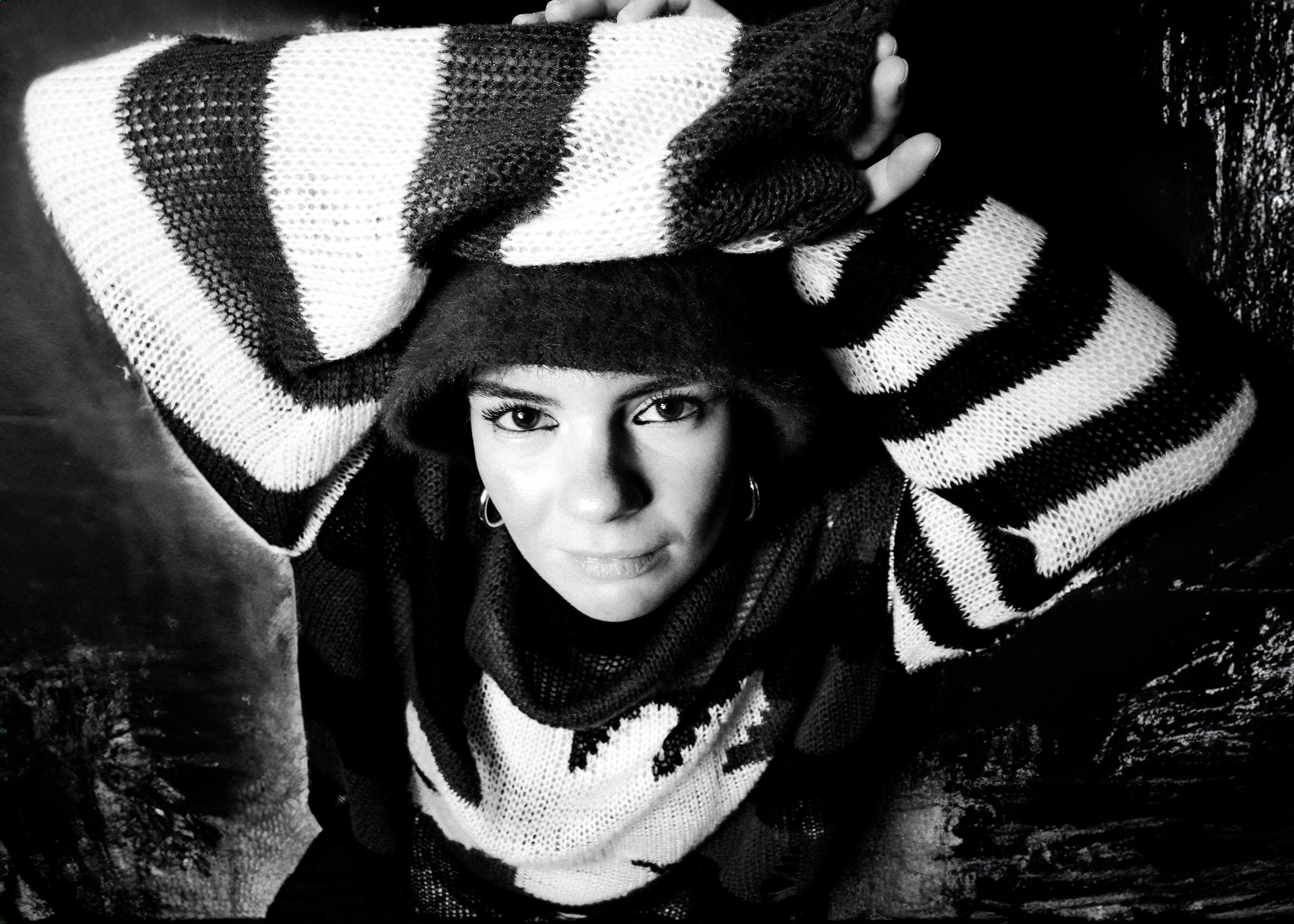 Black and white portrait of woman in striped jumper with hood up, arms raised, looking directly at camera.
