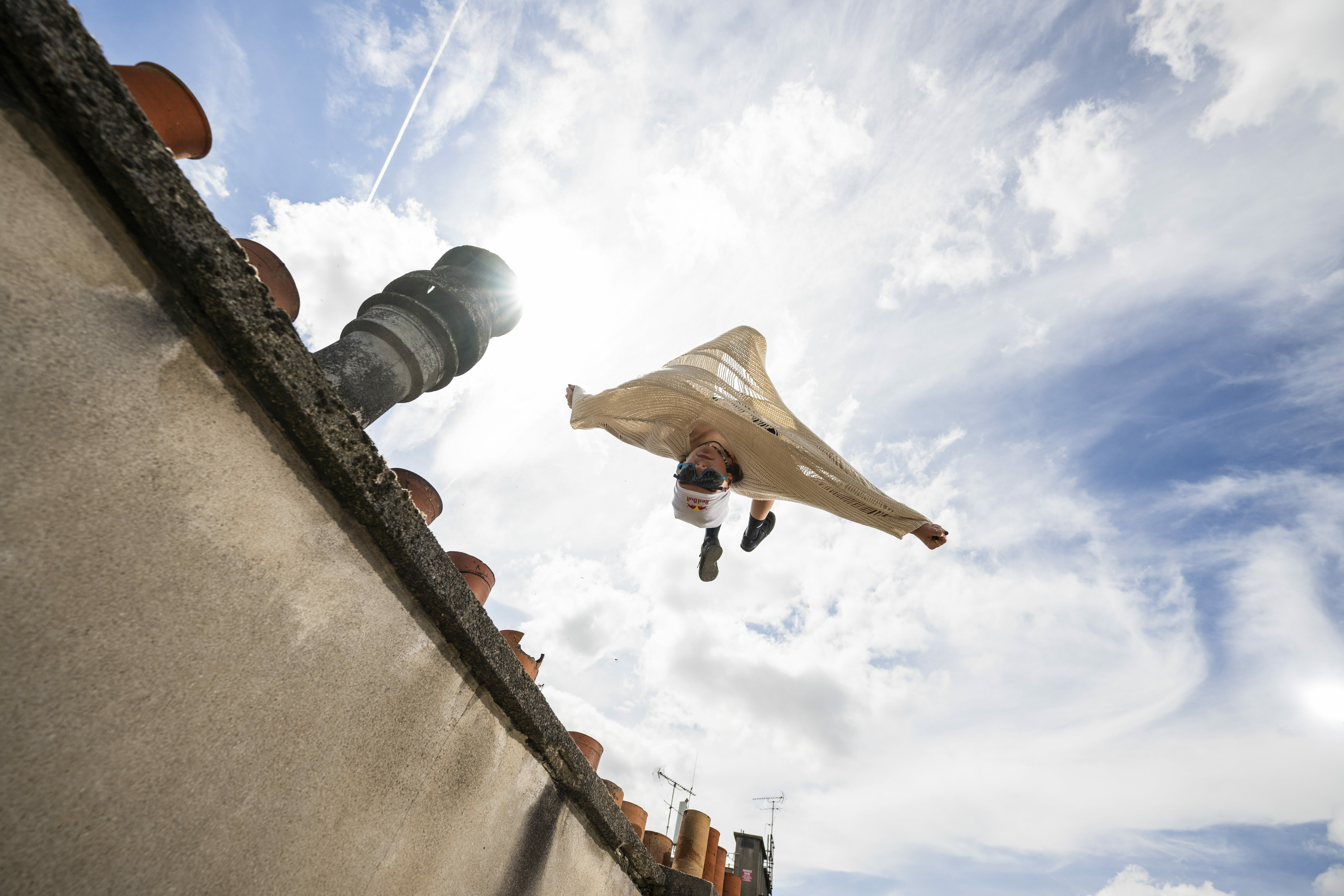 Person hang-gliding above tiled rooftop against cloudy blue sky, captured from low angle looking upward.
