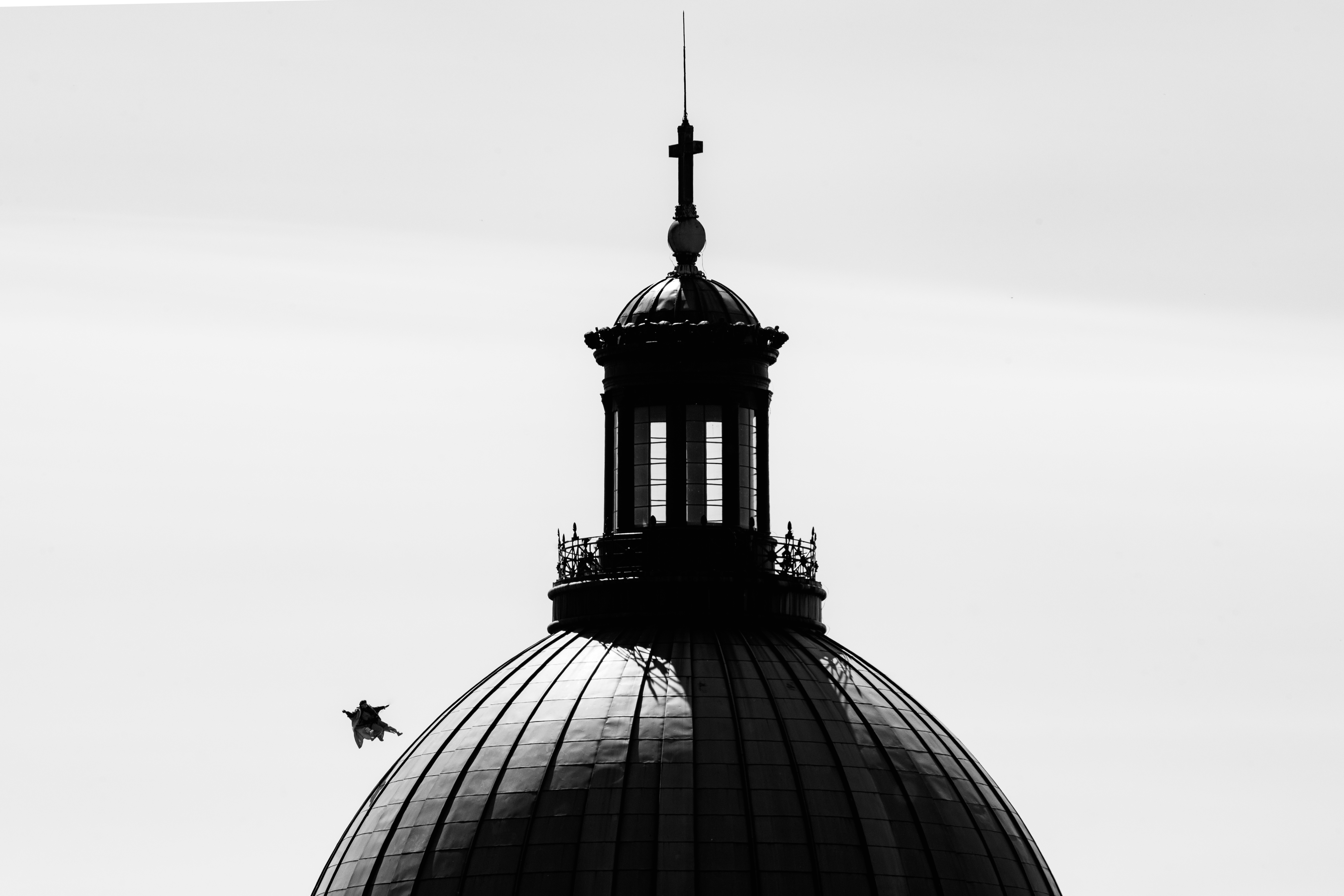 Dark silhouette of ornate church dome with lantern cupola and spire against grey cloudy sky. Small bird flies near the structure.