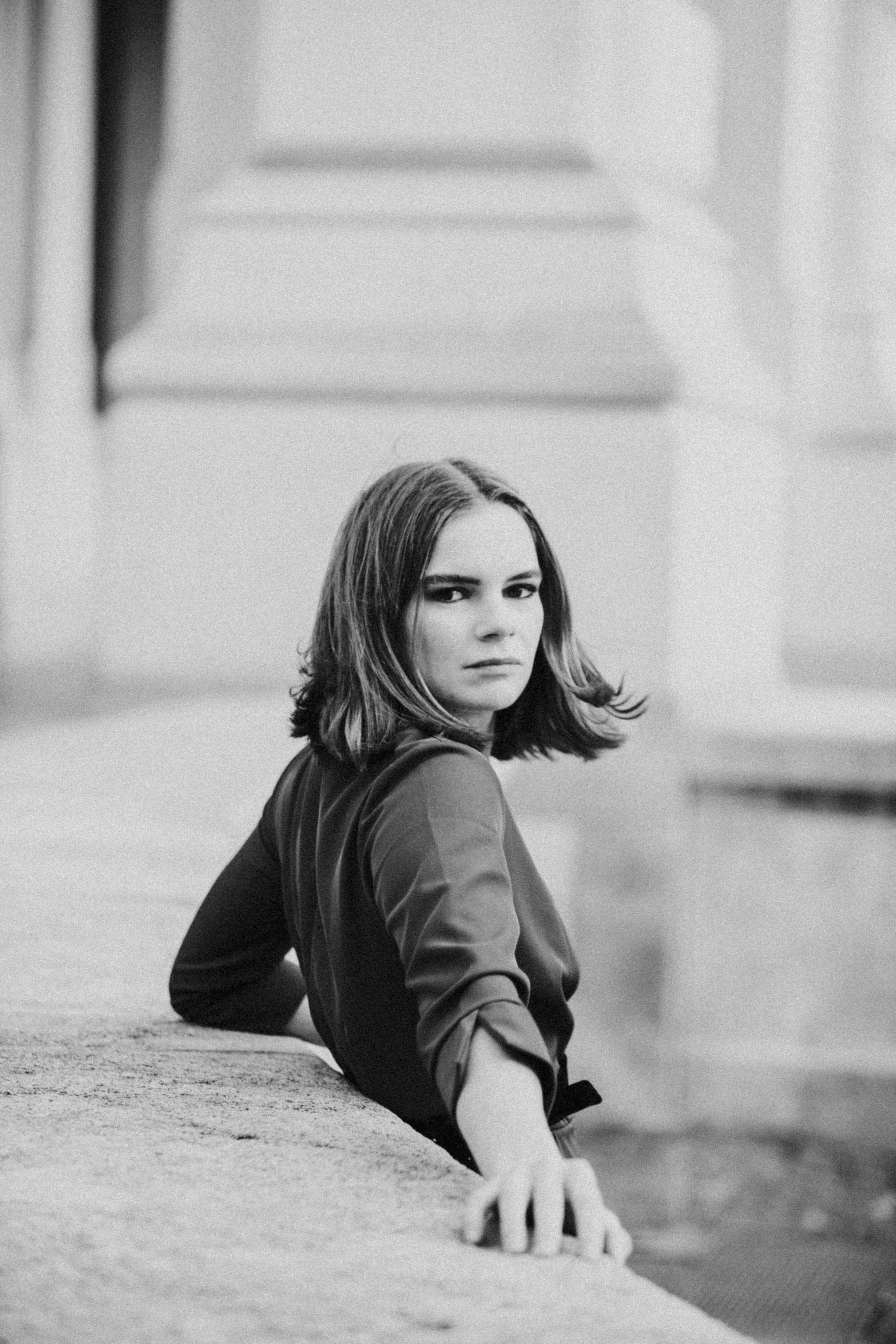 Black and white portrait of woman with shoulder-length hair in dark jacket, leaning on stone surface with blurred architectural background.