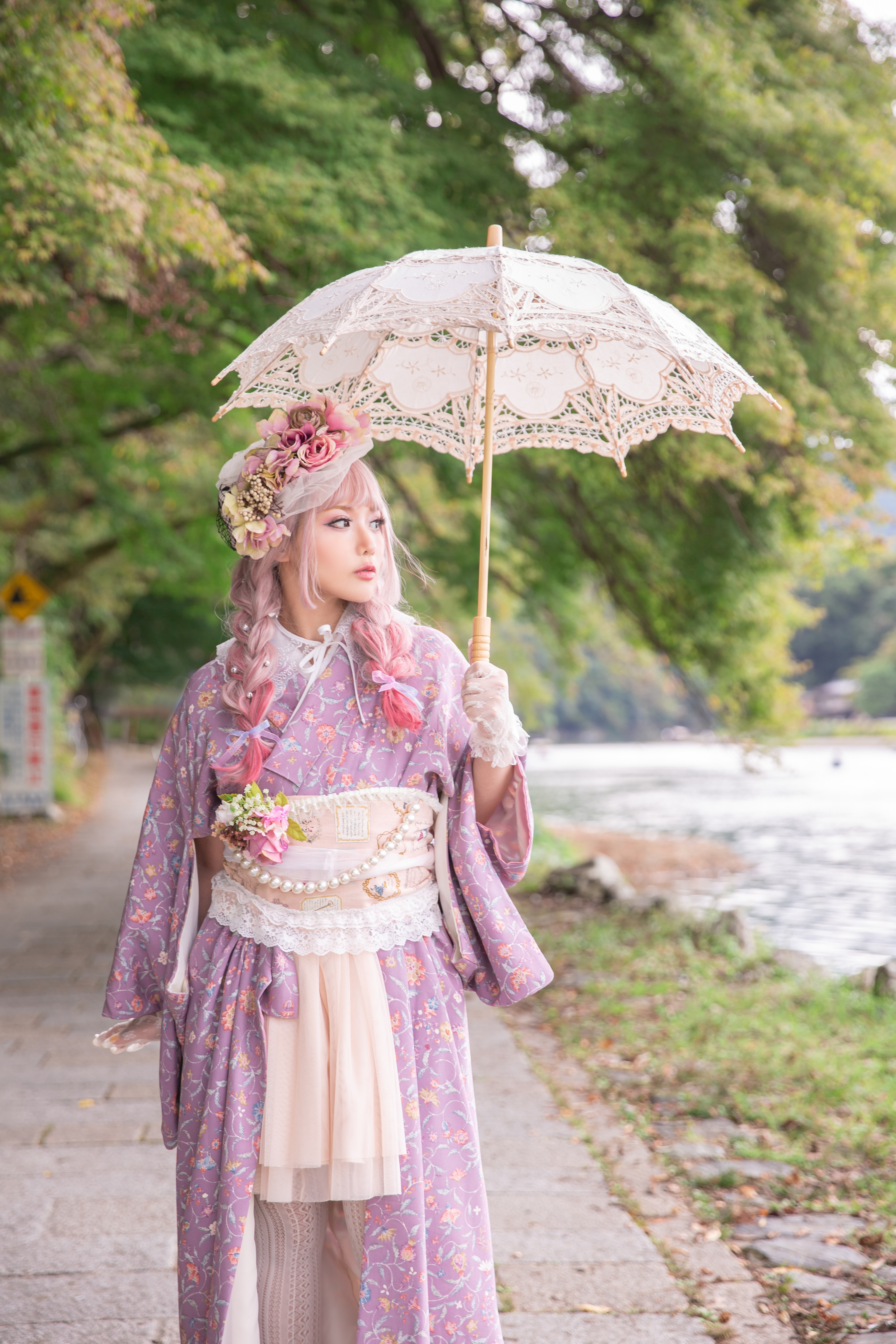 Woman in purple floral dress with cream corset and hair flowers holds white lace parasol on tree-lined path.