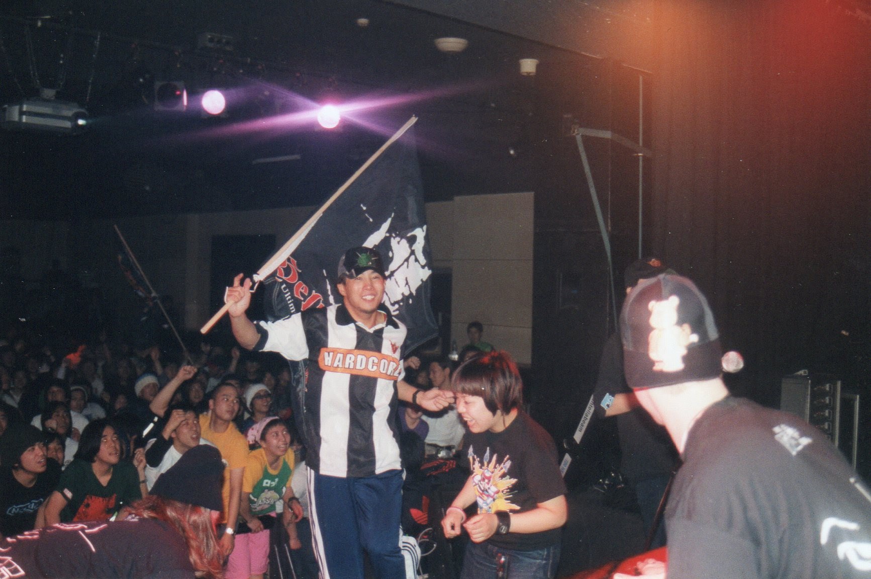 Crowded indoor venue with stage lighting, person in white vest holding microphone above seated audience, purple and orange stage lights visible.