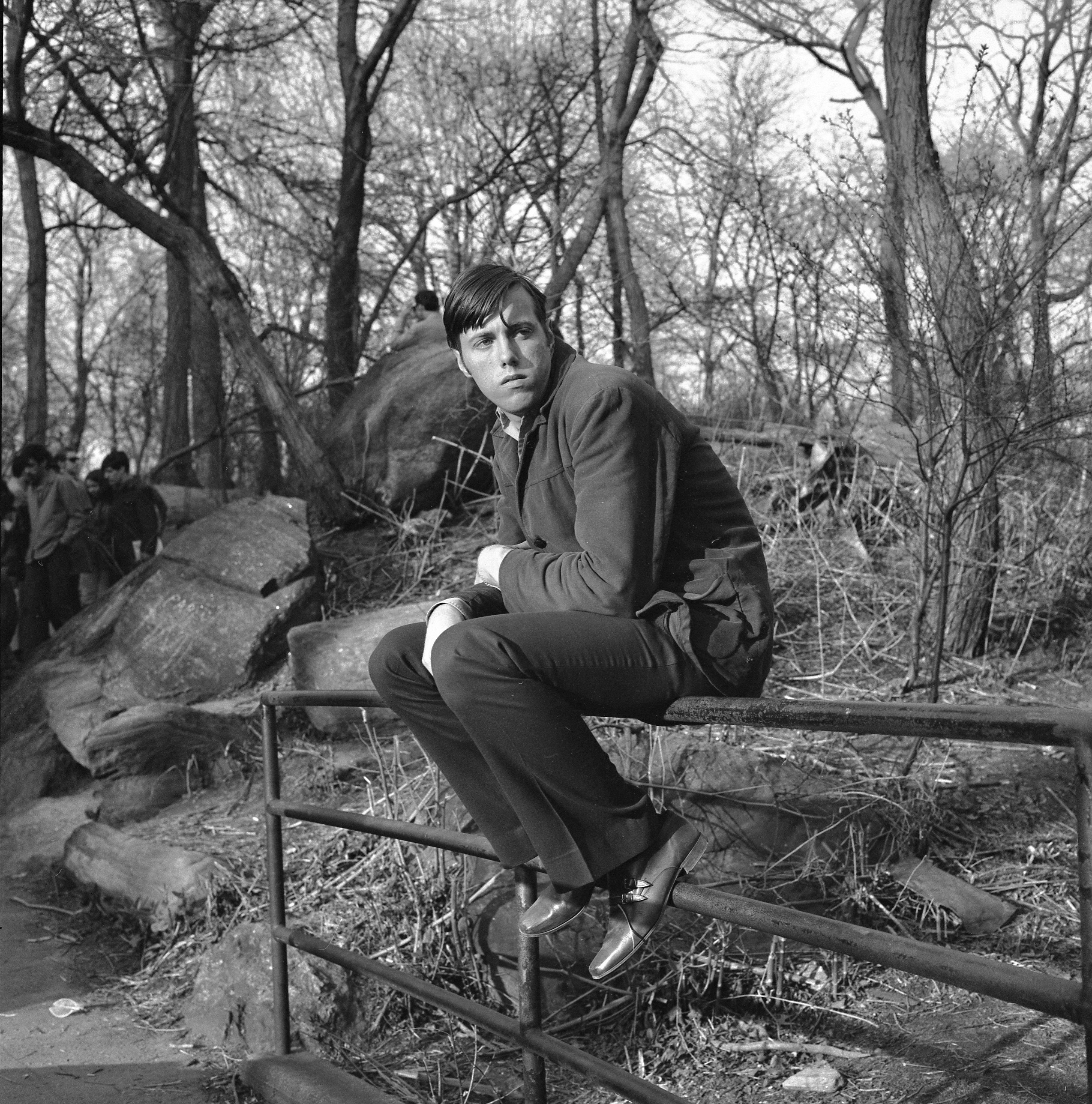 Man in suit sitting on wooden fence rail in wooded area, black and white photograph with bare trees in background.