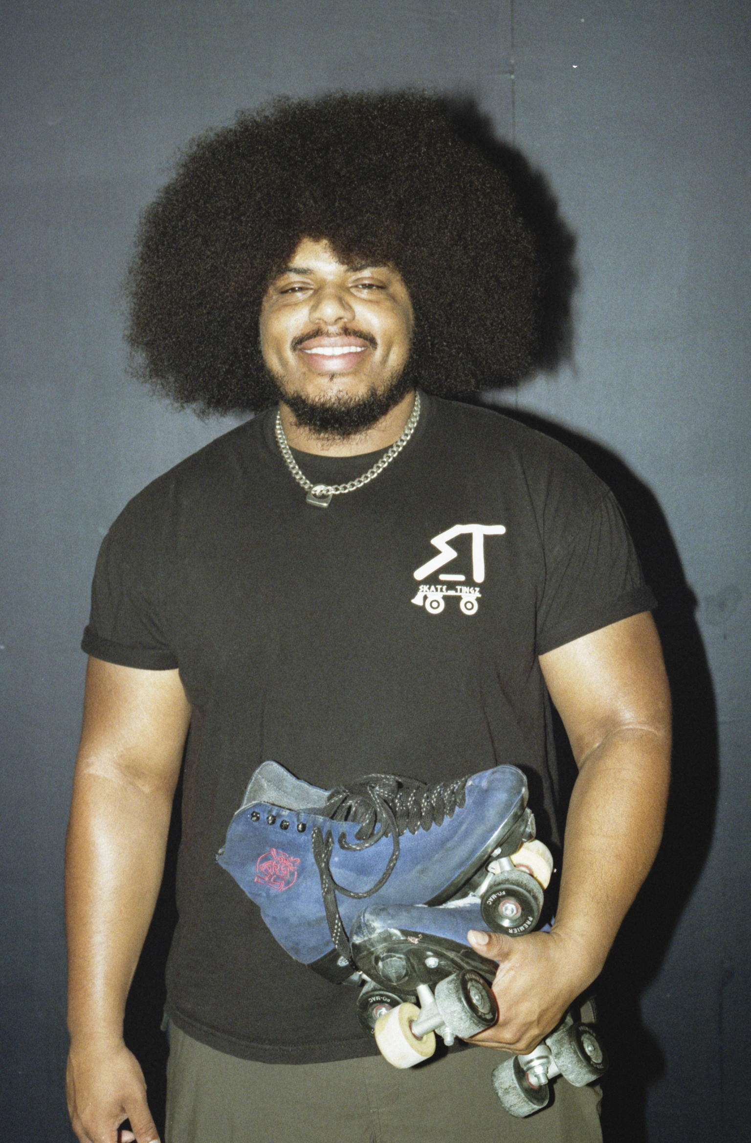 Man with large afro hairstyle wearing black t-shirt and chain necklace, holding roller skates, smiling against grey background.