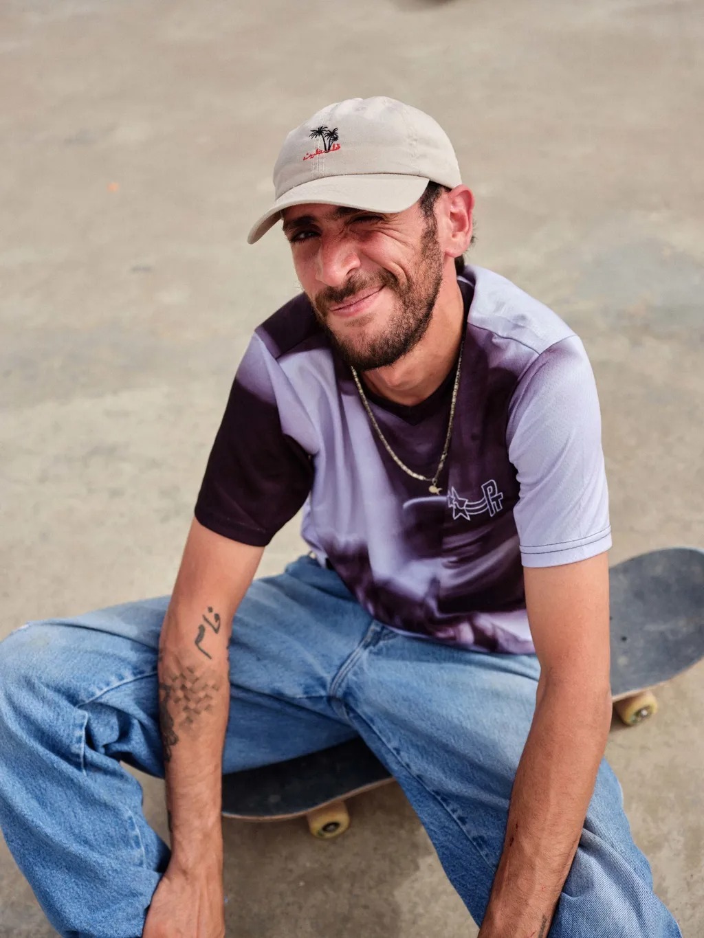 Man with beard wearing white cap and striped purple shirt sitting on skateboard in blue jeans, smiling at camera outdoors.