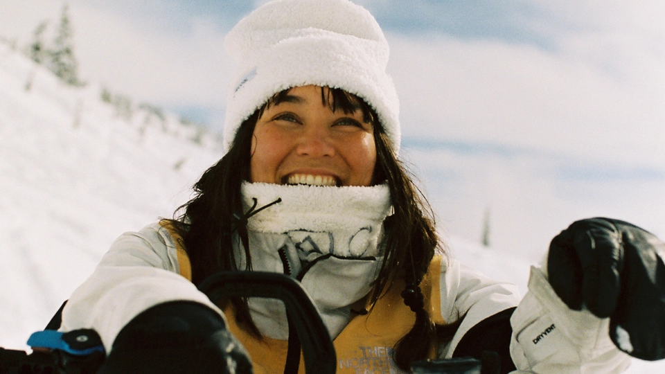 Smiling person in white winter hat and jacket with face covering, wearing black gloves, snowy mountain landscape background.