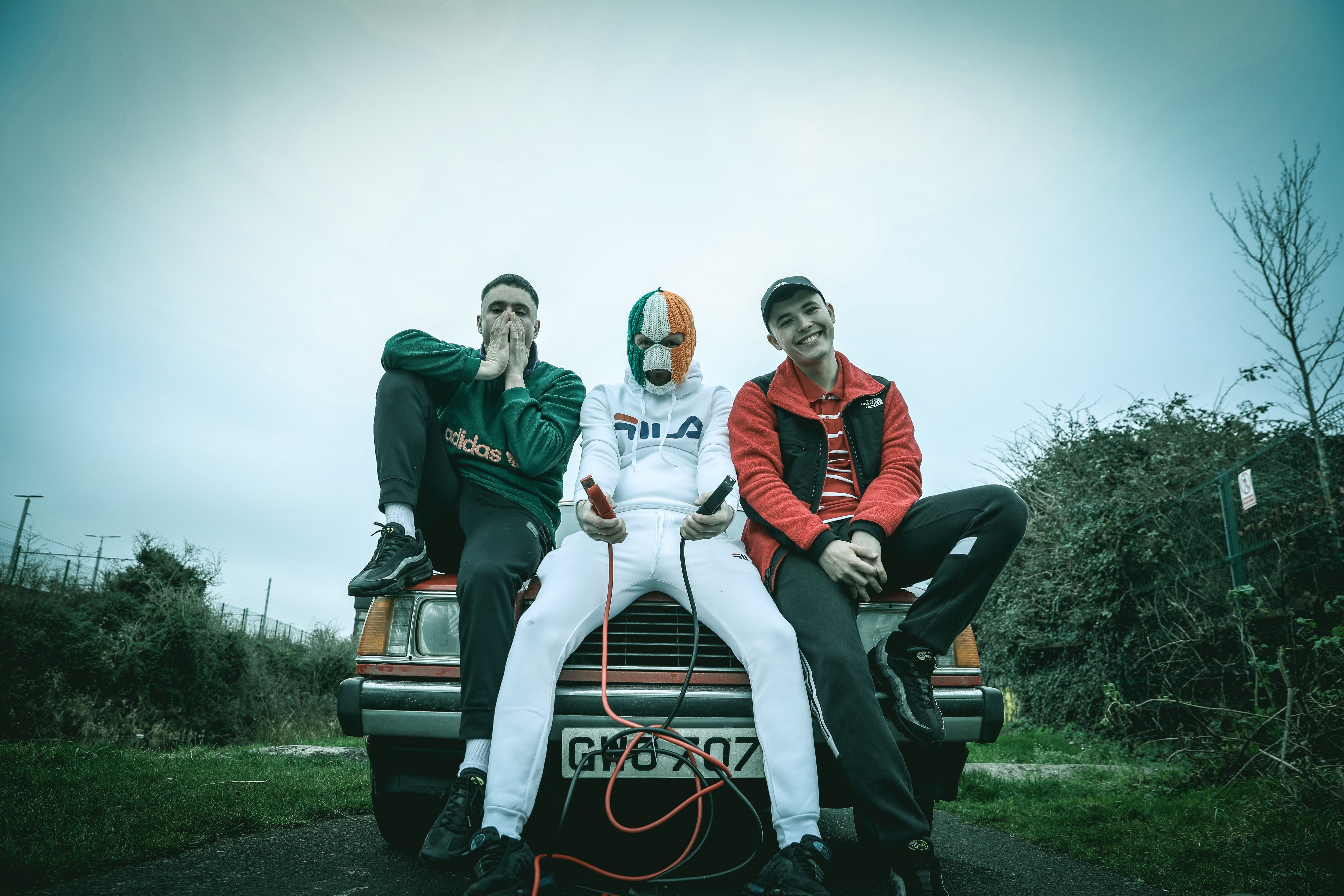 Three people wearing masks sit on brown car bonnet against pale blue sky. Left figure in black, centre in white with orange/white mask, right in red jacket.