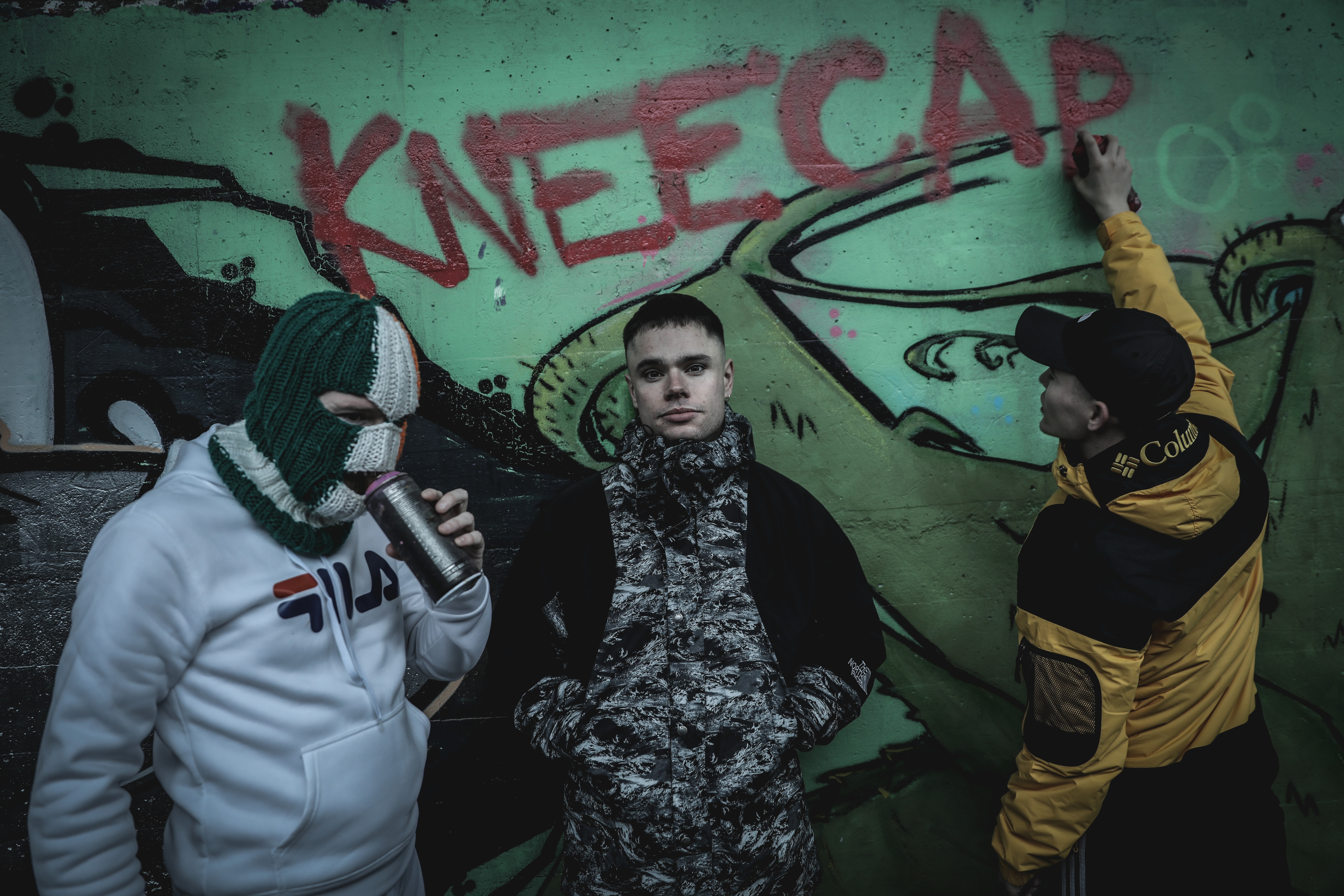 Graffitied urban wall with three individuals dressed in street wear, one holding a spray can.