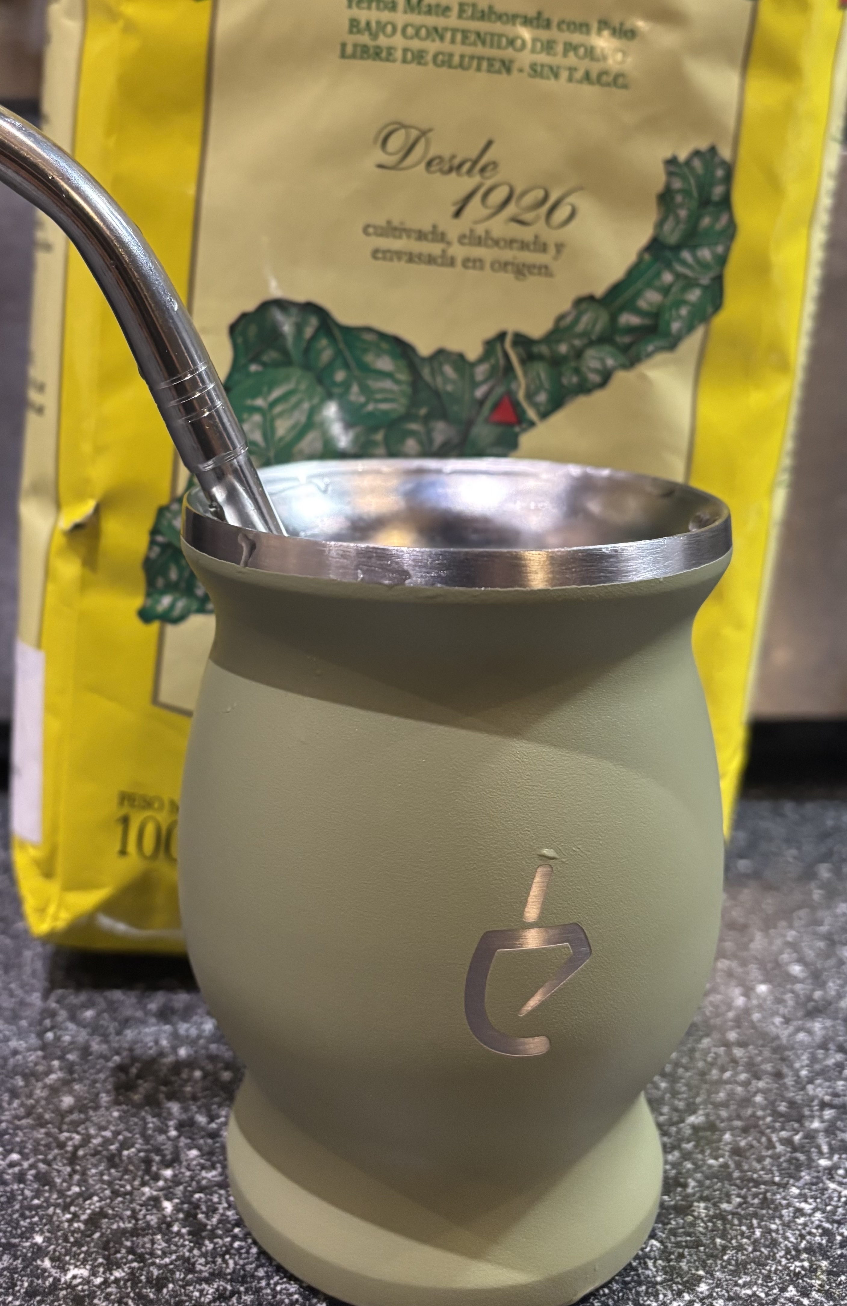 Cylindrical glass mug with olive green surface and metallic rim, placed in front of a yellow and green food packaging with text "Desde 1926" (Since 1926).