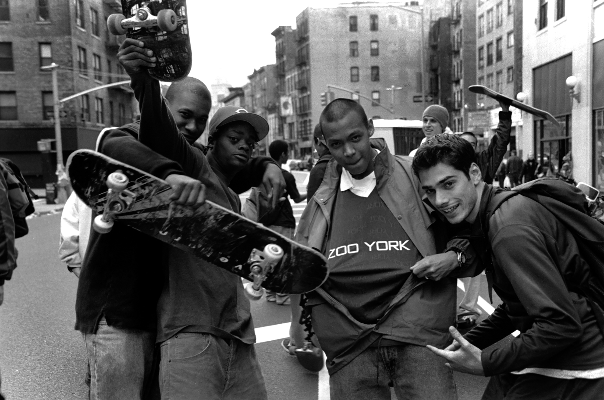 Black and white image of four young men on city street, one holding skateboard above head, others gathered around smiling at camera.