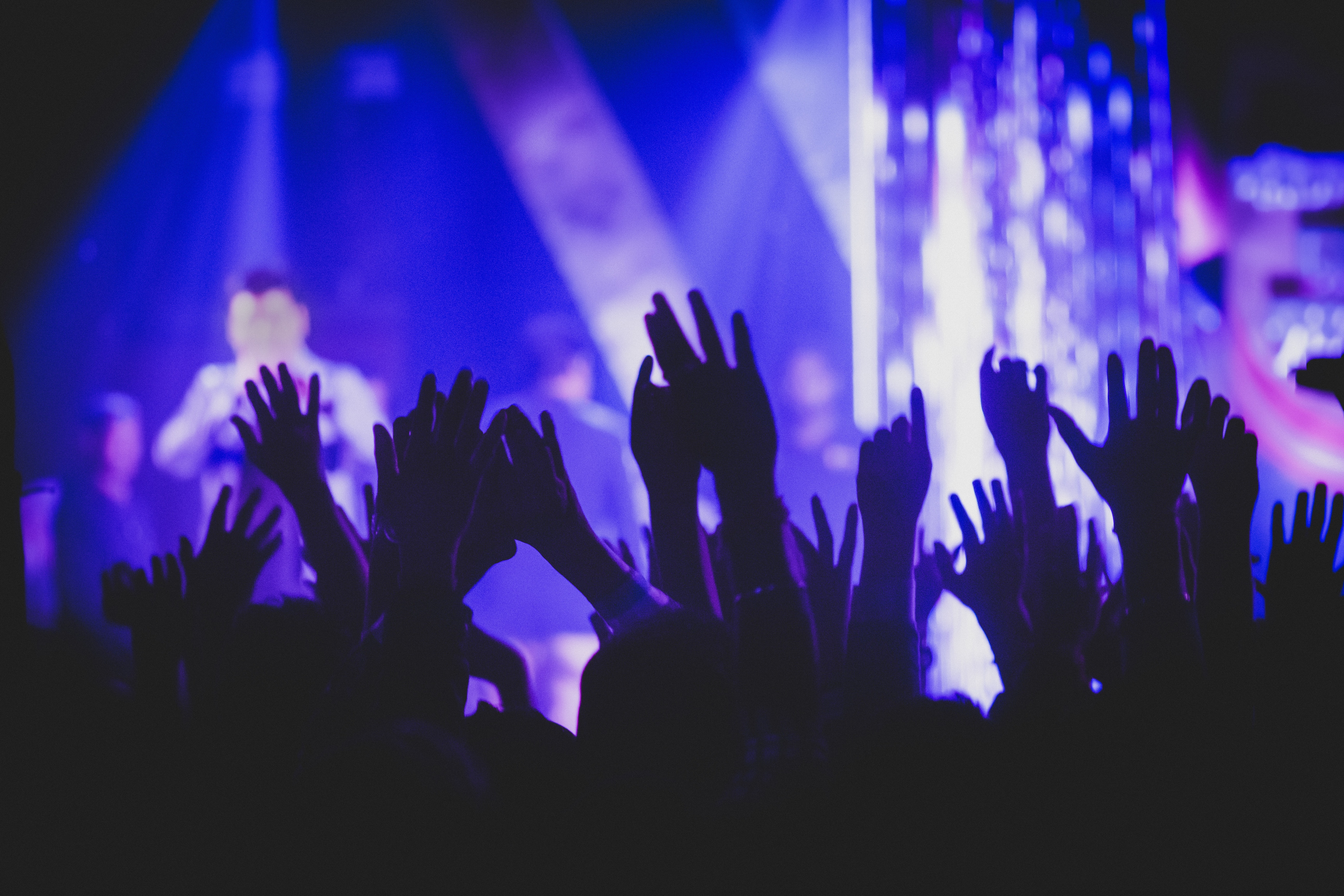 Silhouetted crowd with raised hands against bright blue and purple stage lighting beams cutting through darkness.