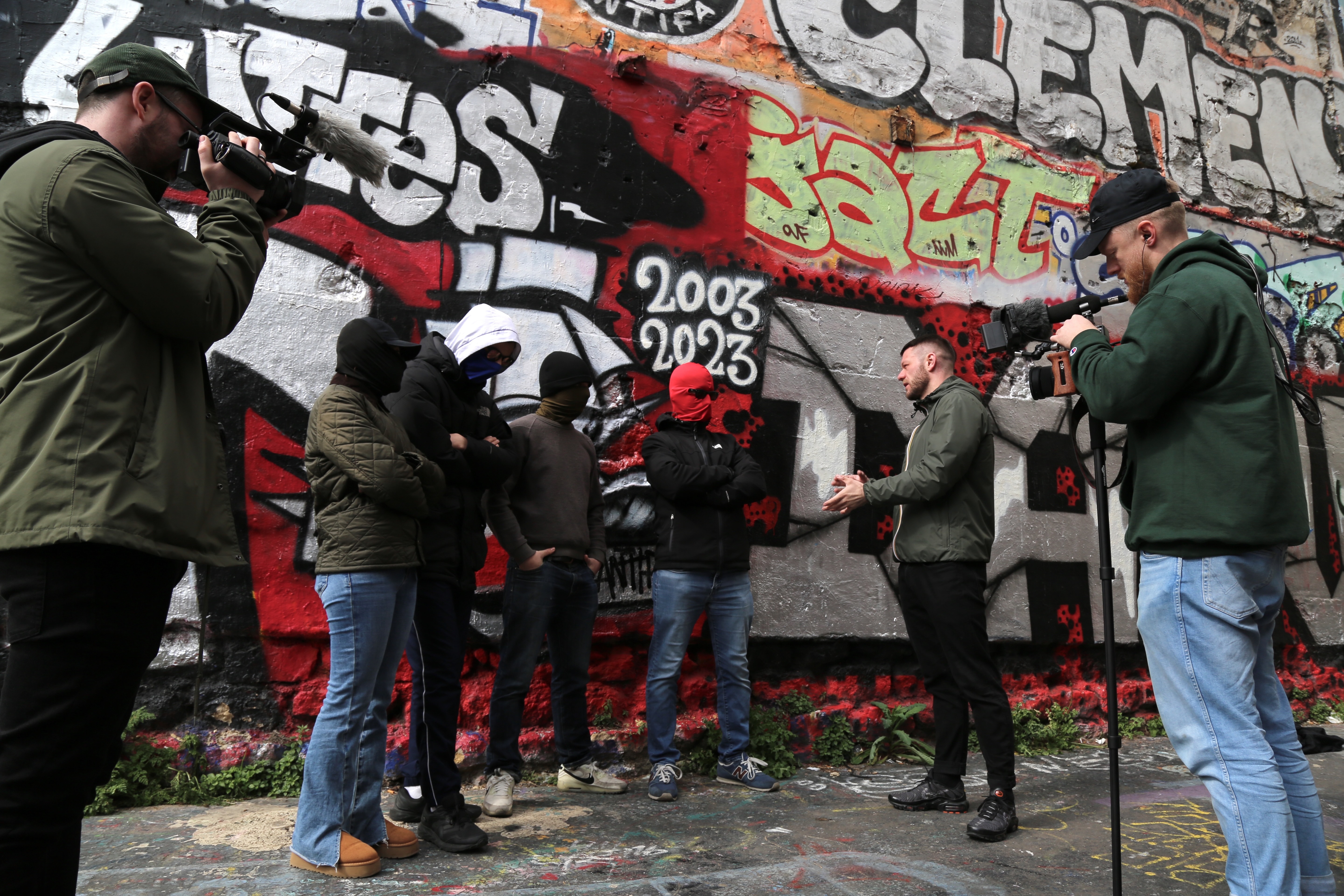 Film crew with cameras recording people spray-painting graffiti on colourful wall featuring red, black, and white street art.