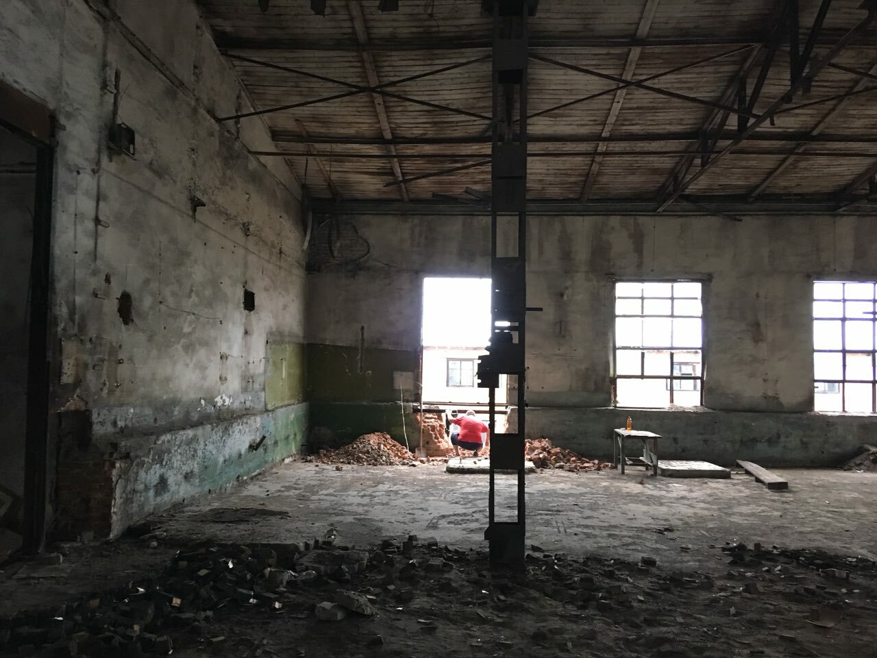Dilapidated interior of an abandoned industrial building with damaged walls, a ruined floor, and large windows letting in natural light.