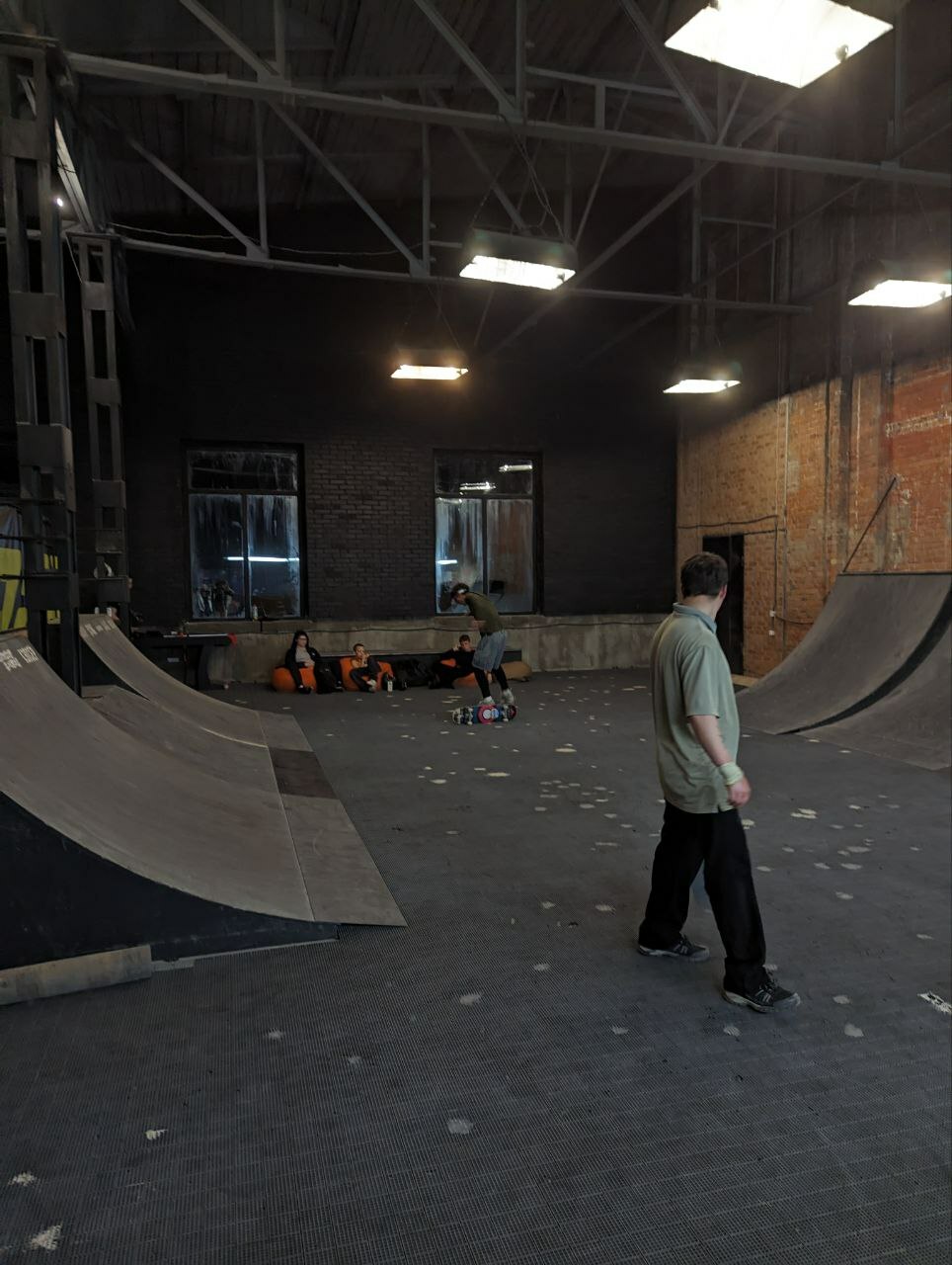 A skate park with ramps and equipment, lit by overhead lights. A person is standing in the foreground, facing away from the camera.