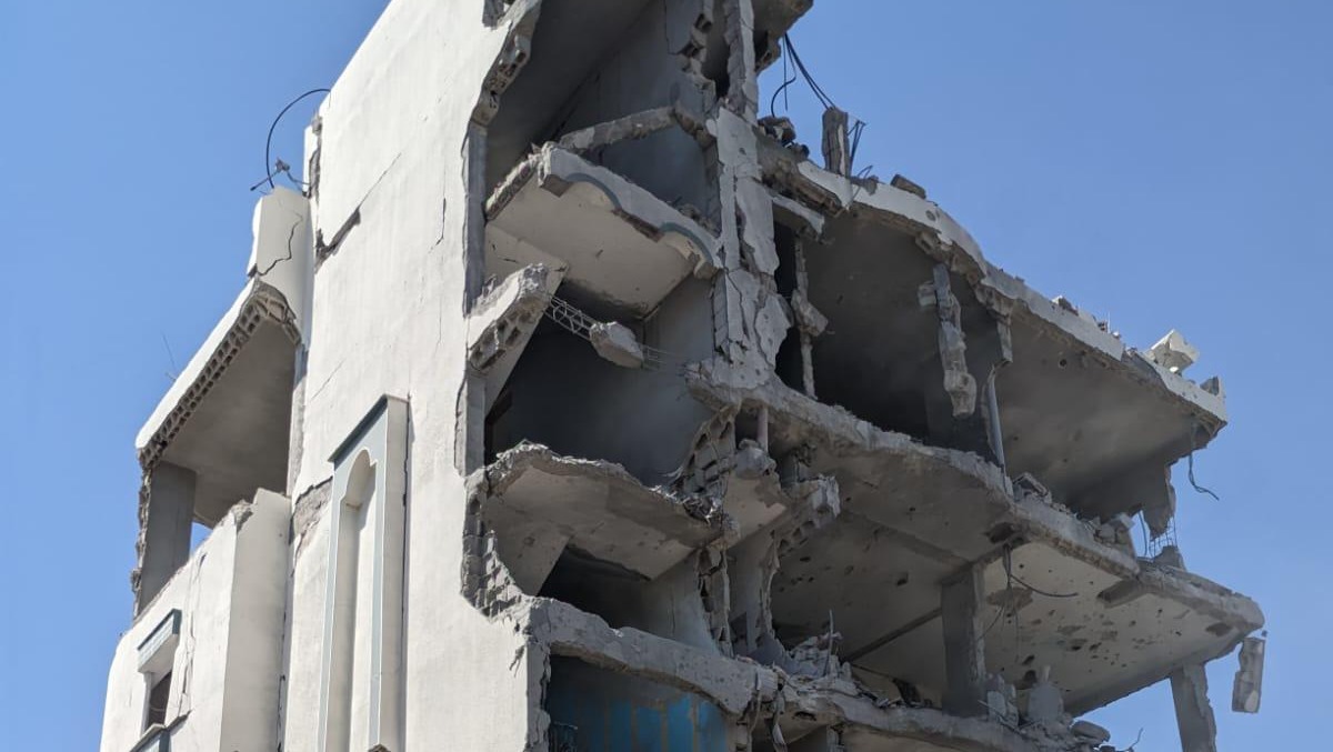 Severely damaged multi-storey building with collapsed concrete floors, exposed rebar, and crumbling white walls against blue sky.