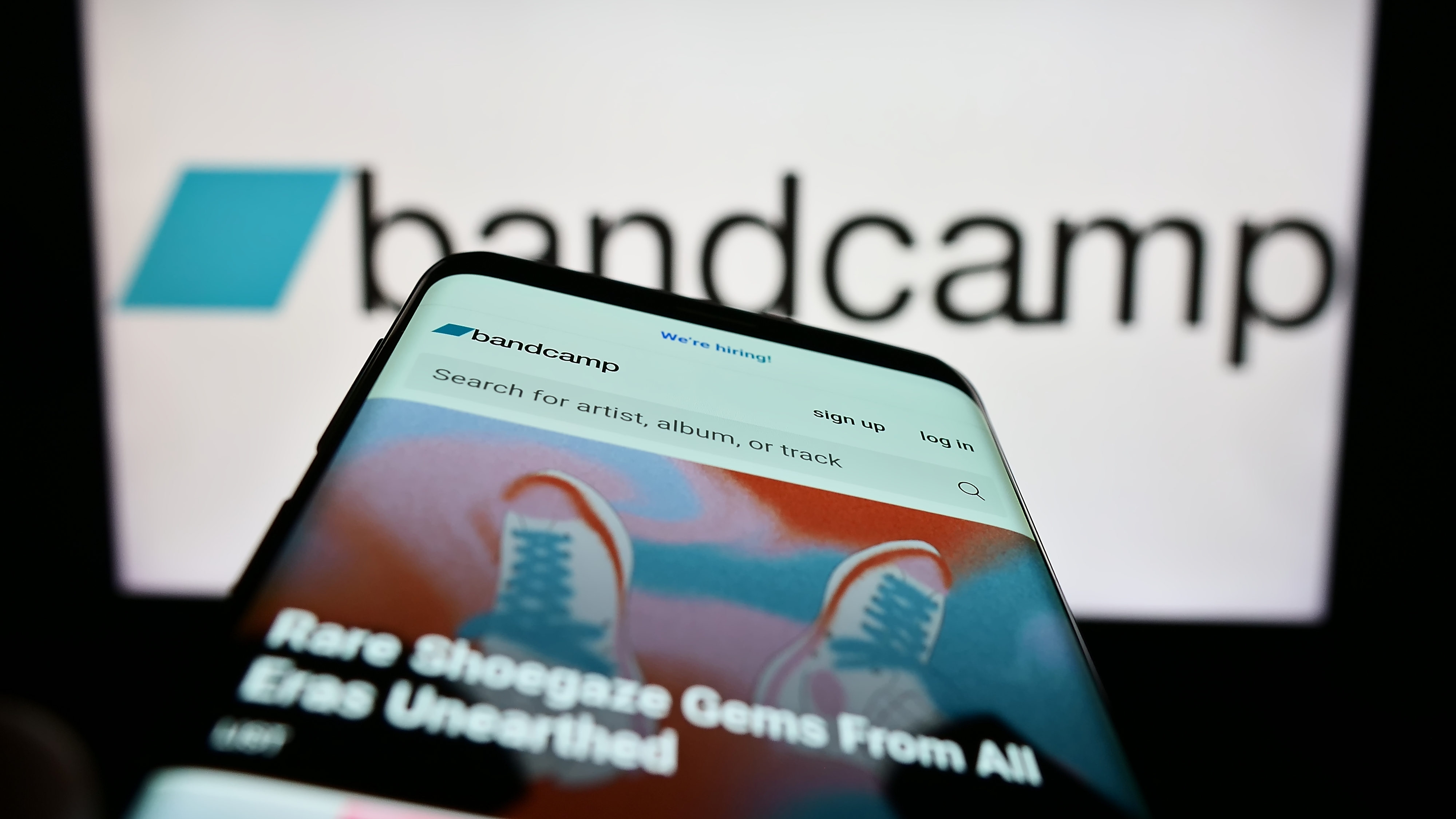 Smartphone displaying Bandcamp website with colourful album artwork, positioned in front of blurred Bandcamp logo on computer screen.