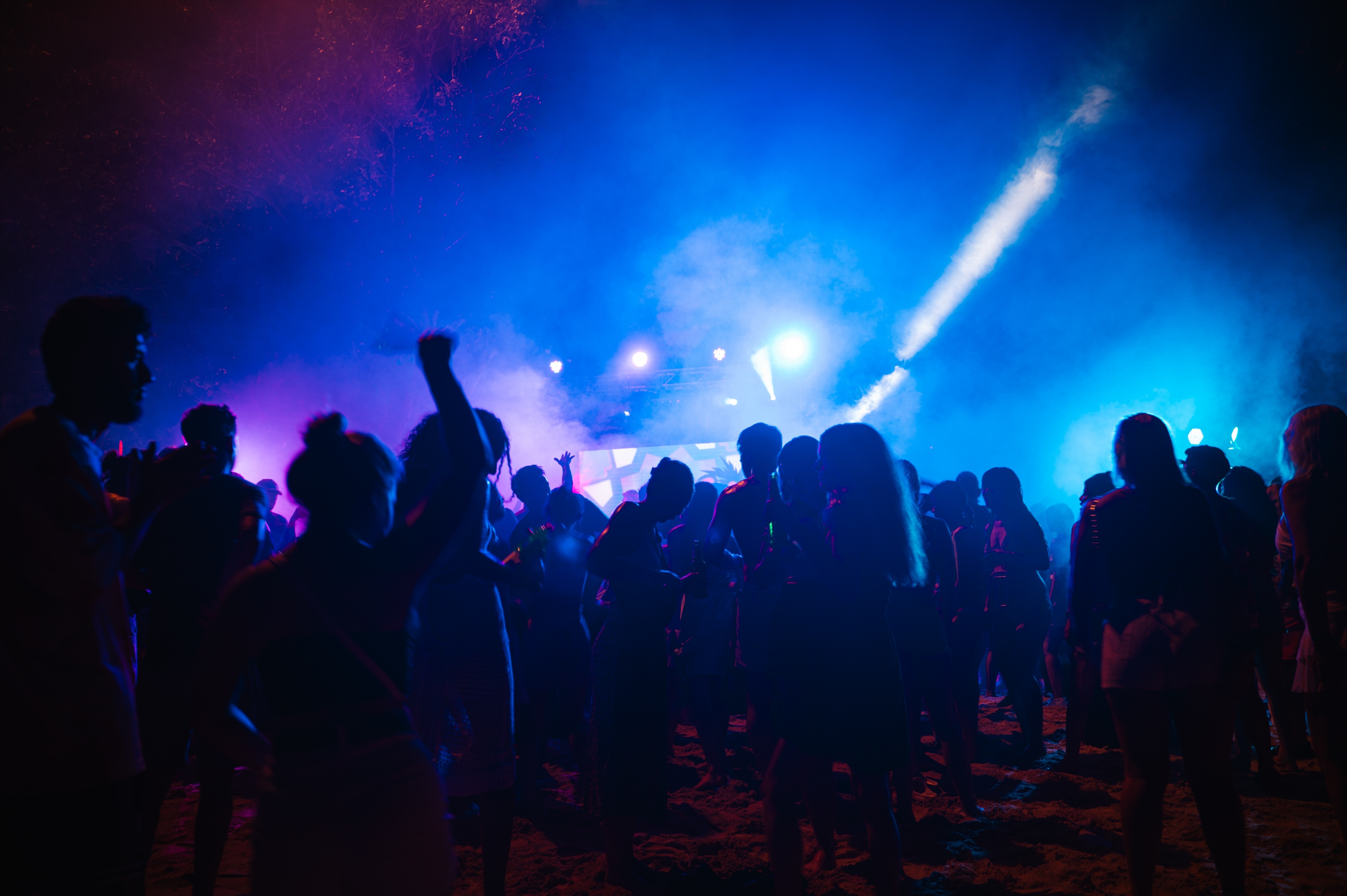 Crowd of silhouetted people at a nighttime event with colourful lighting and a bright spotlight on stage.