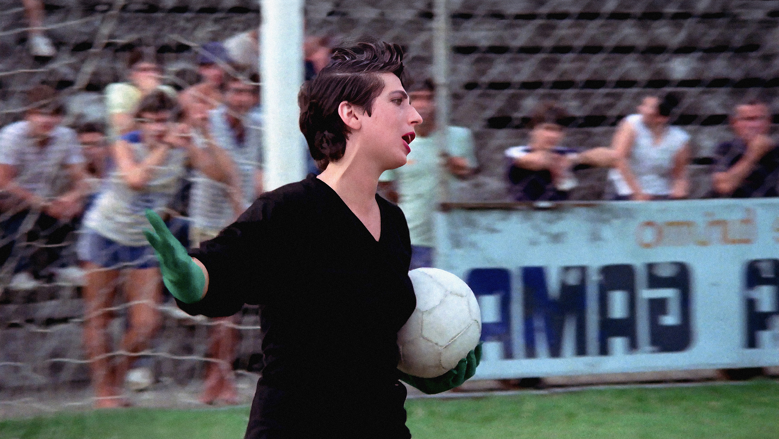 Man in black t-shirt walking on a football pitch, holding a ball.