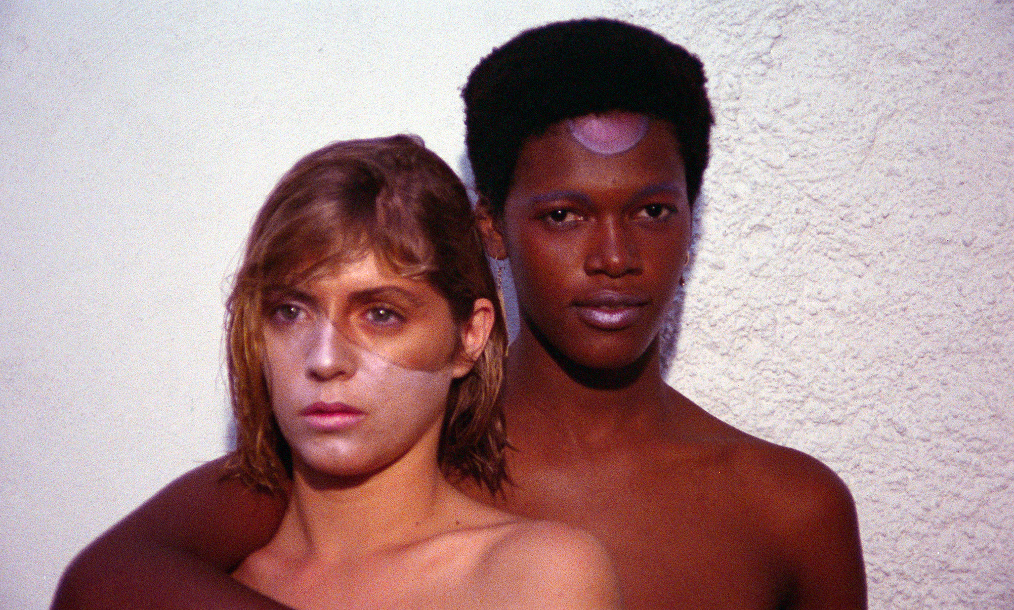 Two individuals, a woman with long brown hair and a man with dark skin, standing close together against a plain white background.