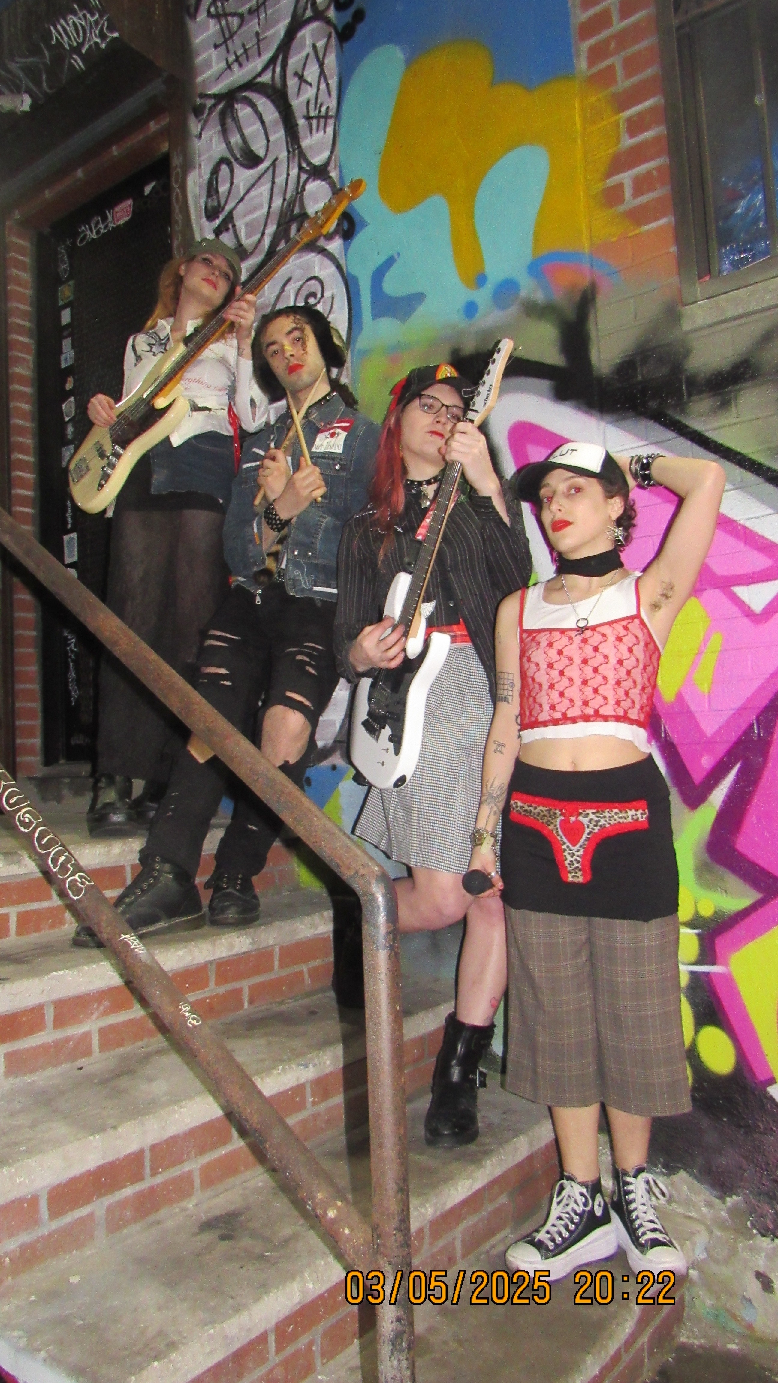 Group of young adults in edgy, colourful outfits posing with guitars in front of graffiti-covered wall.