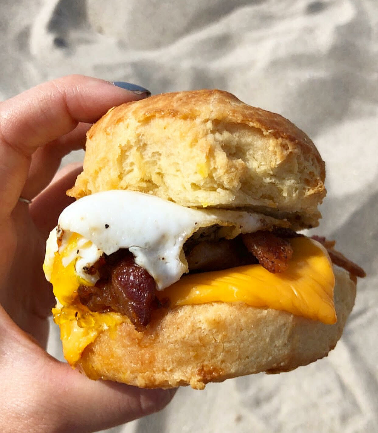 A hand holding a breakfast sandwich with a golden biscuit, a fried egg, melted cheese, and a meat patty.