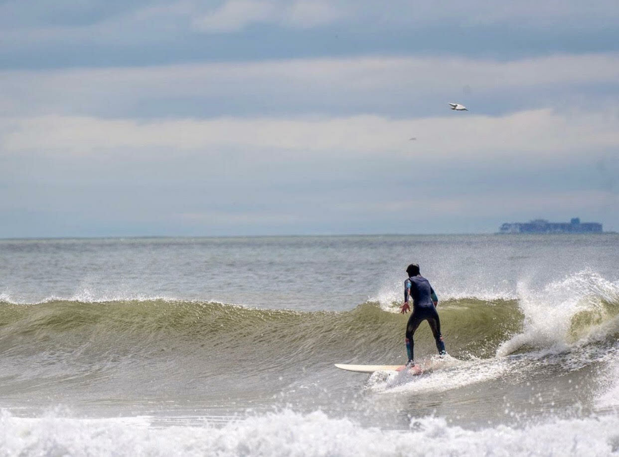 Surfer riding wave, distant land visible, plane overhead, cloudy sky.