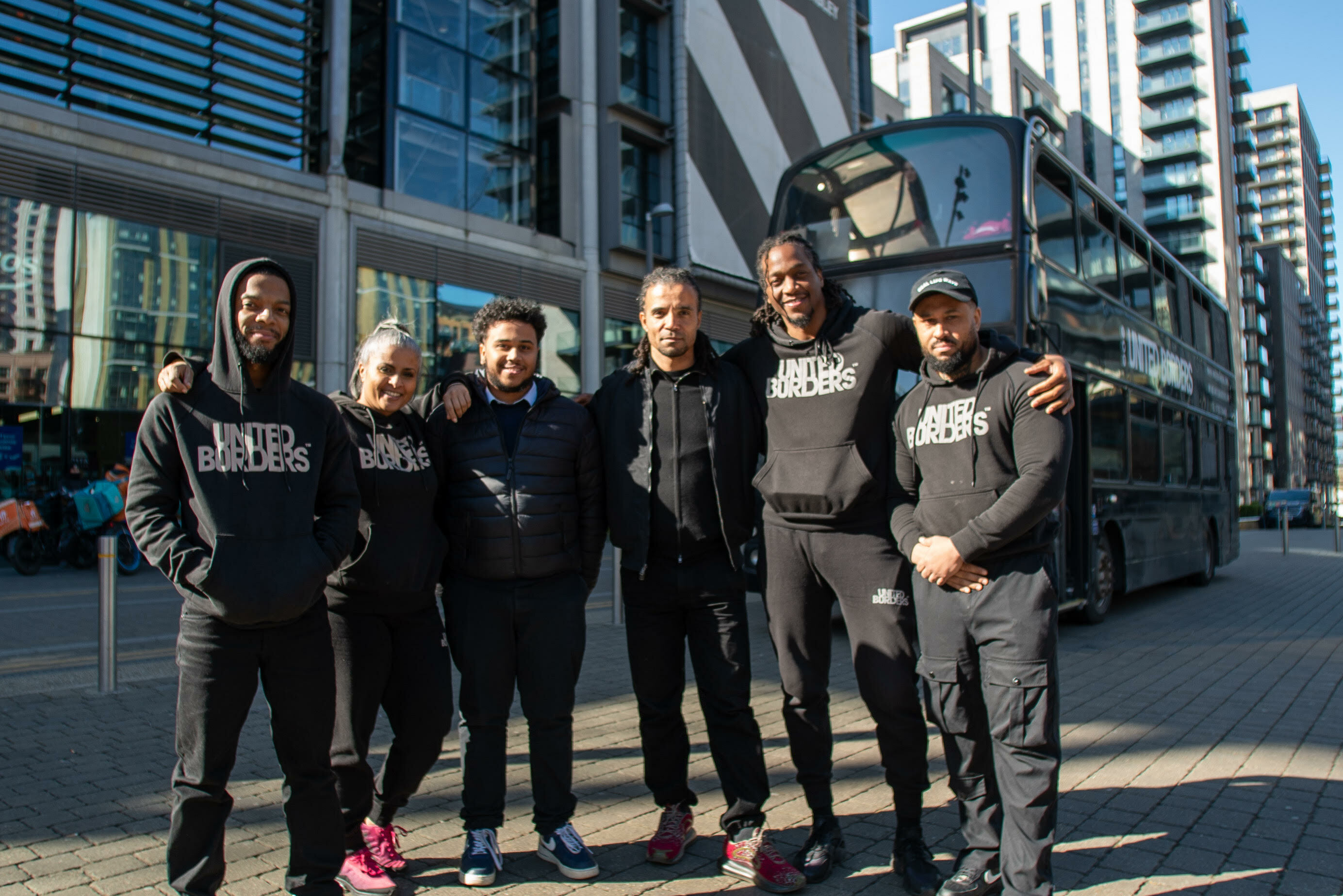 Group of 6 people wearing black and grey hoodies with "OUTSIDERS" written on them, standing outside a modern glass building.