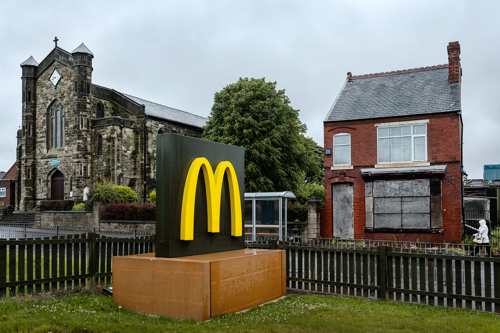 McDonald's golden arches logo on brown plinth beside Gothic stone church and red brick house with wooden fence under cloudy sky.