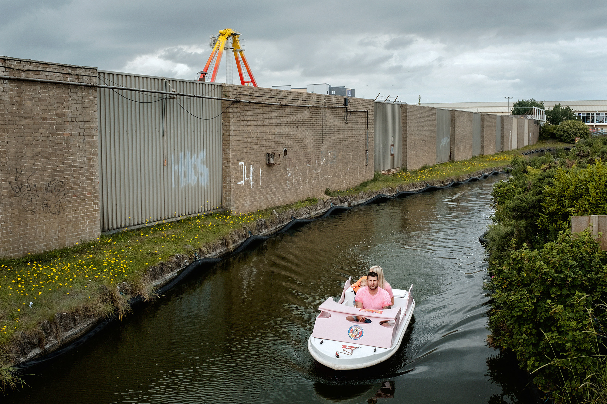 Small white boat with two people on narrow canal beside concrete industrial wall with orange crane structure visible above.