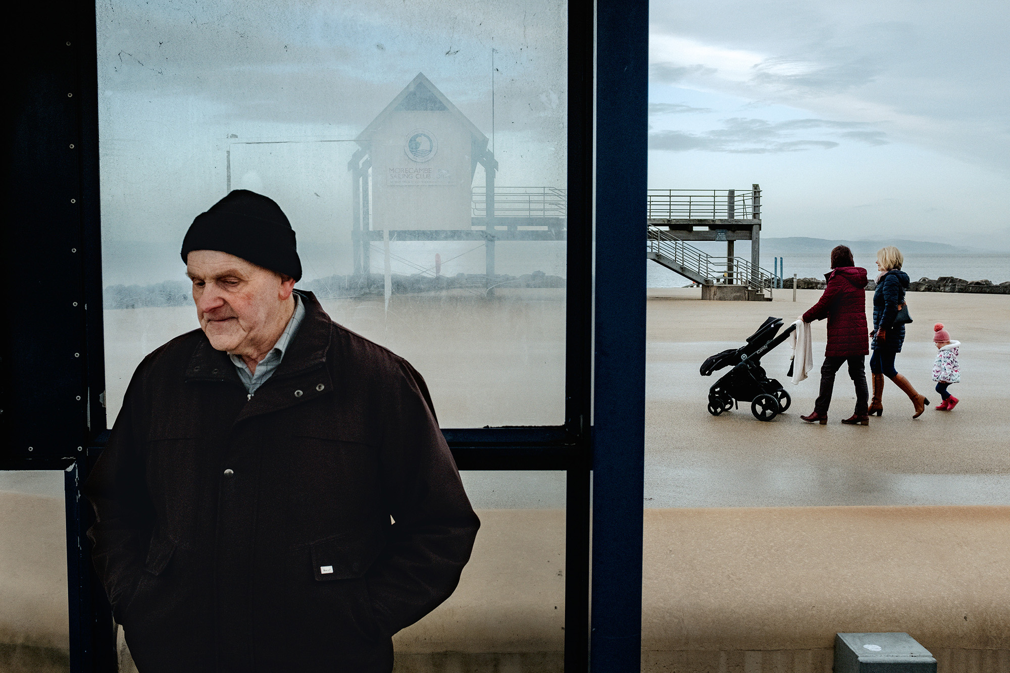 Elderly man in black coat and beanie sits inside glass shelter whilst family with pram walks on beach behind him.