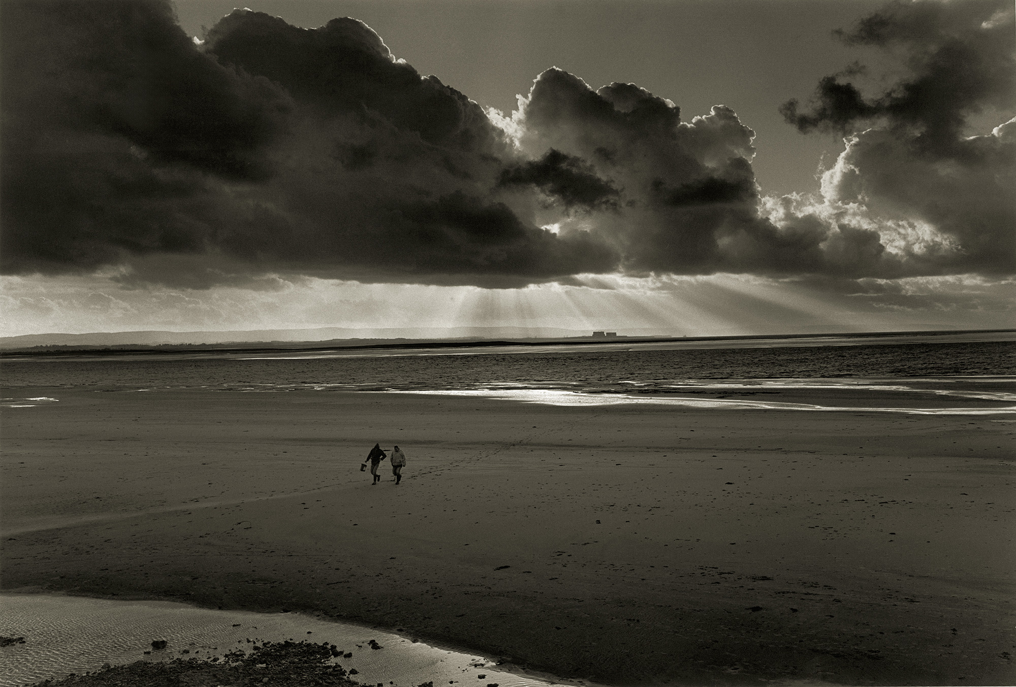 Two figures on expansive beach with dramatic dark clouds overhead, light breaking through storm clouds, wet sand reflecting sky.