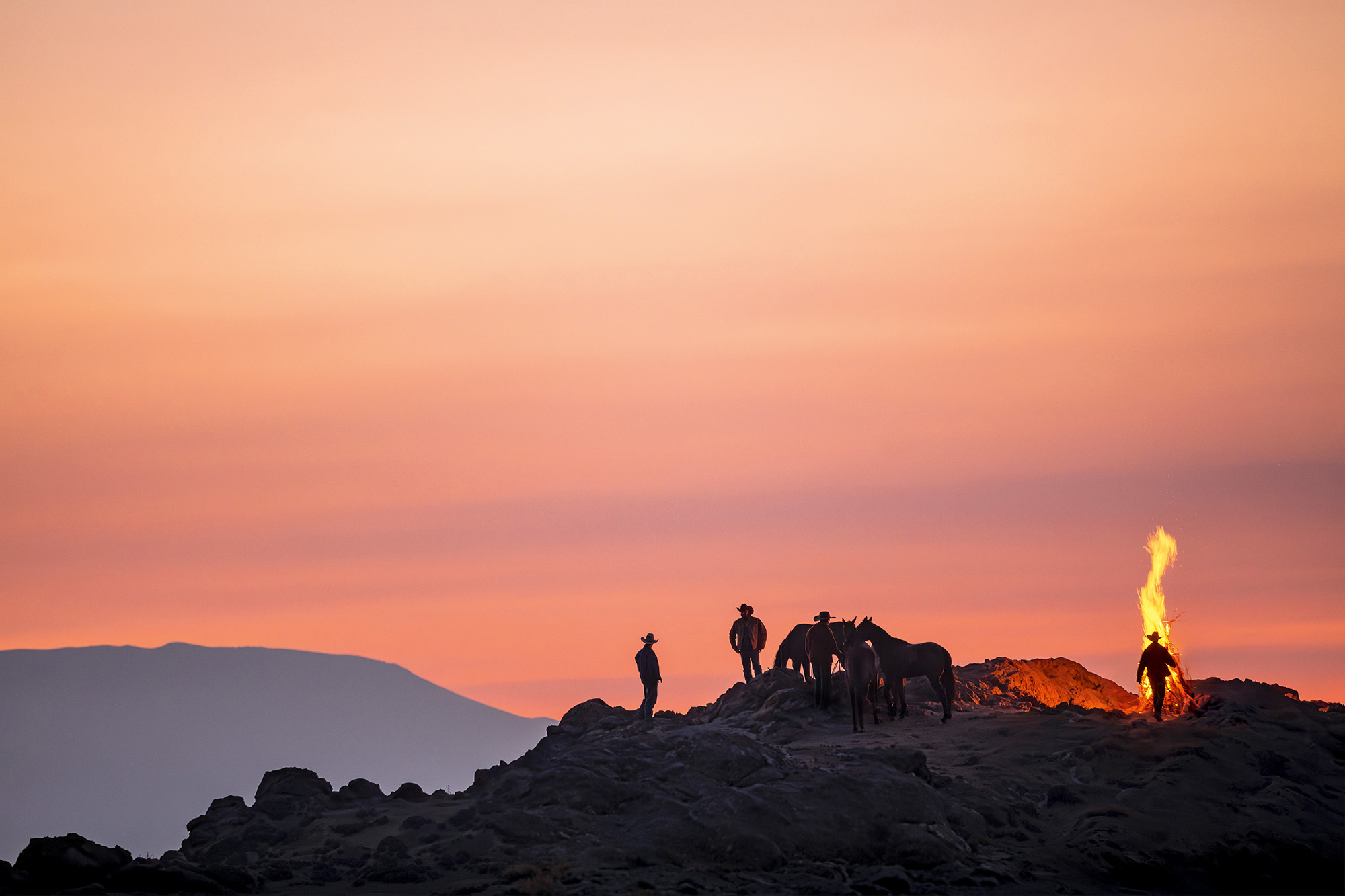 Silhouettes of people and vehicle on rocky ridge against pink-orange sunset sky with distant mountains visible.