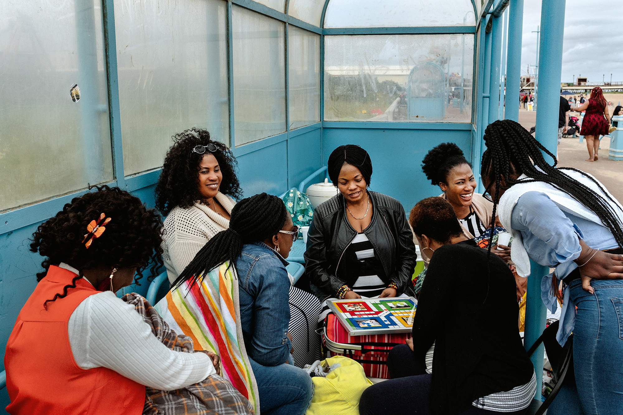 Group of women sitting and standing in blue-walled glass enclosure, some reading books, wearing casual clothing in various colours.