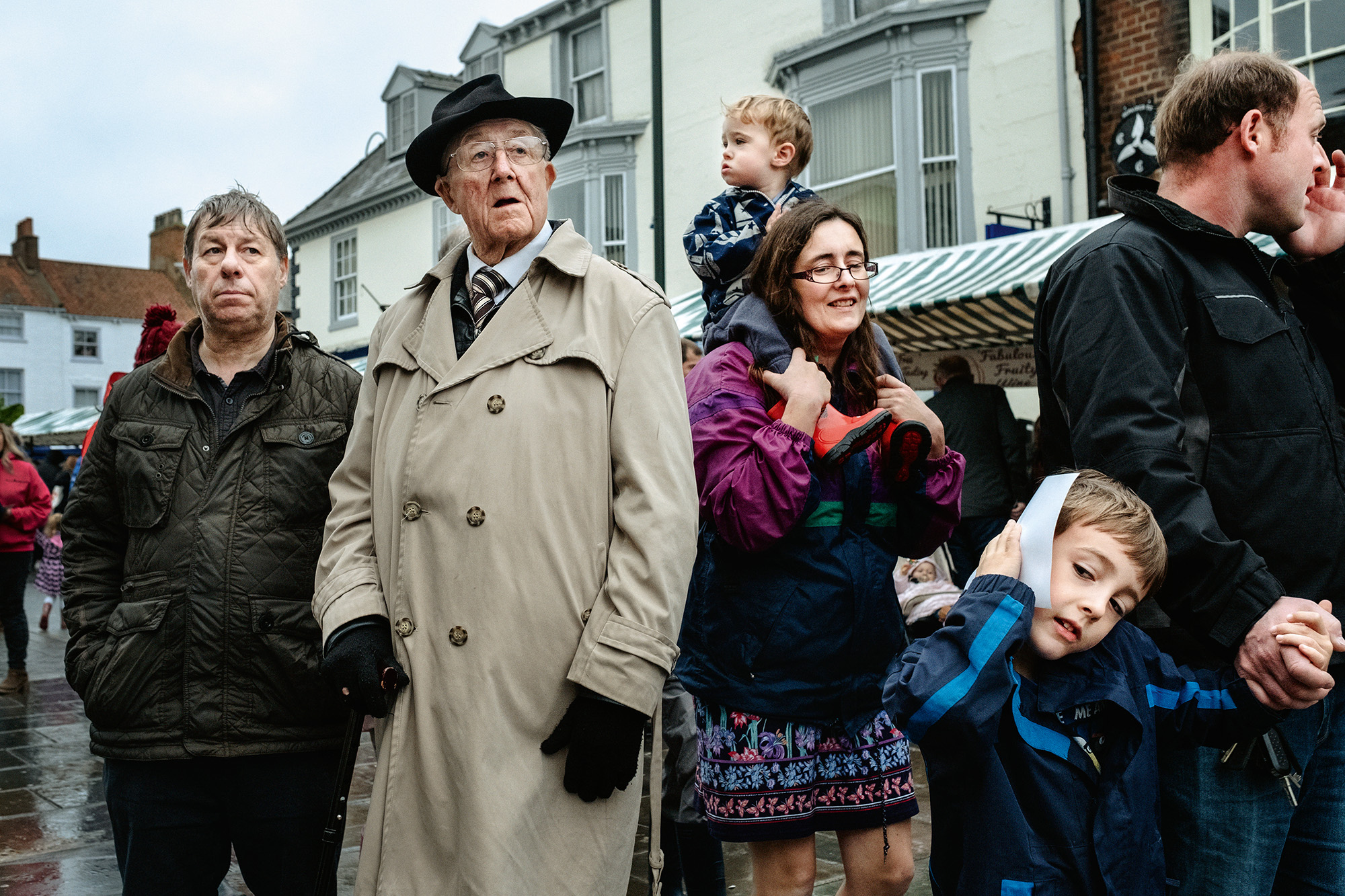 Group of people gathered on street, including elderly man in beige coat and hat, families with children, against backdrop of white Georgian buildings.
