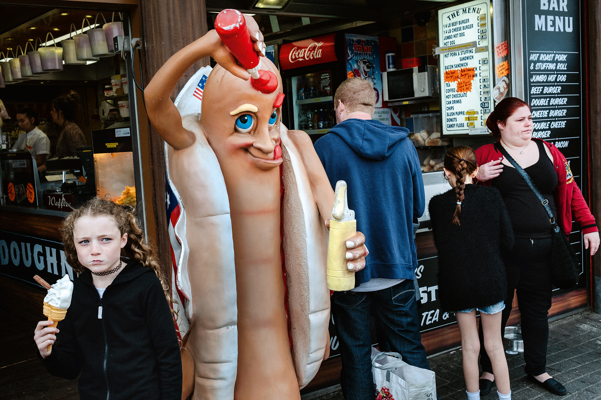 Person in hot dog costume with red cap and blue eyes stands outside food stall, with child and adults nearby.