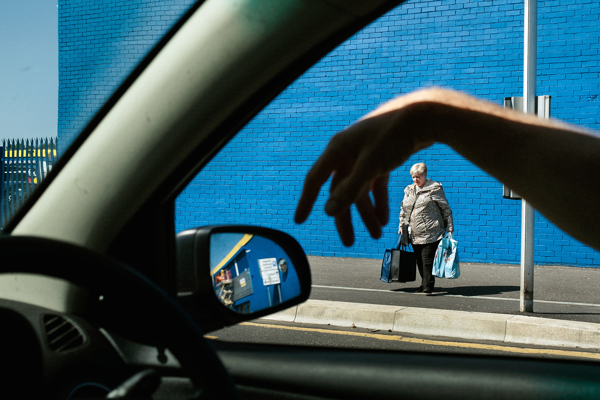 View through car window showing elderly person with shopping bags walking past bright blue tiled wall, steering wheel and wing mirror visible.