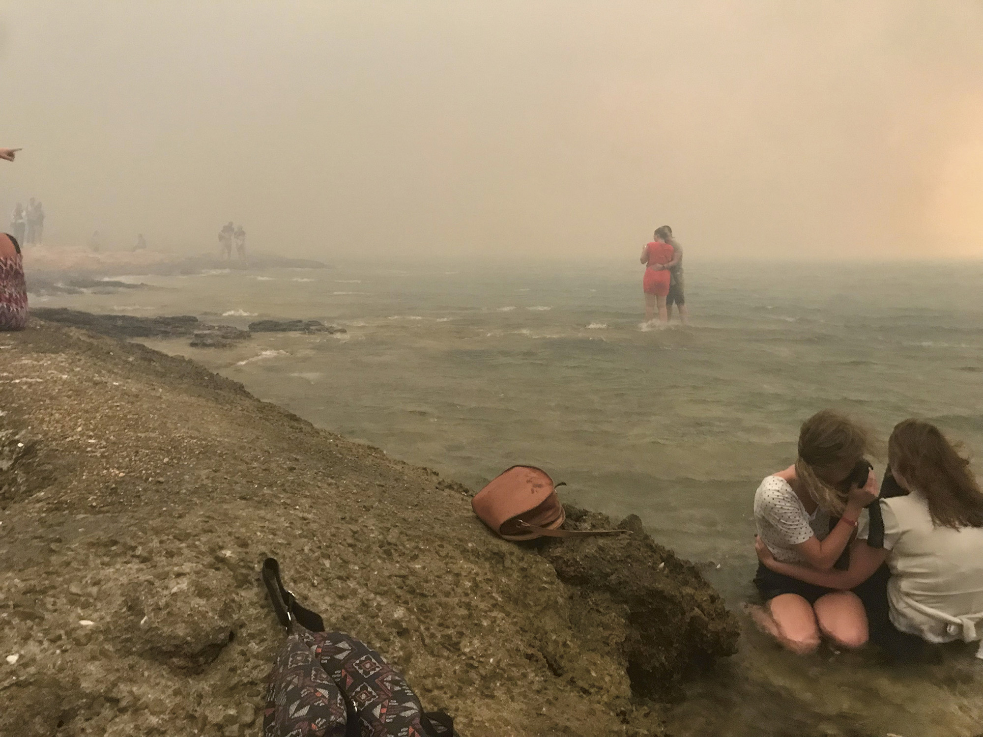 Beach scene with orange-tinted smoky sky. Two children digging in sand in foreground, adults and objects scattered along shoreline in hazy distance.