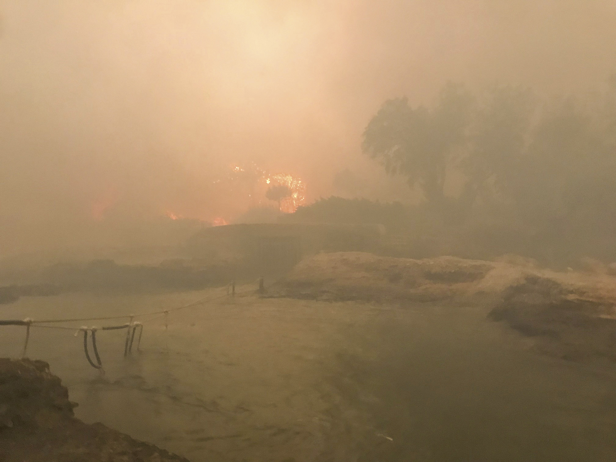 Hazy orange and pink sky with thick smoke obscuring sun over rural landscape with fields and farm structures.
