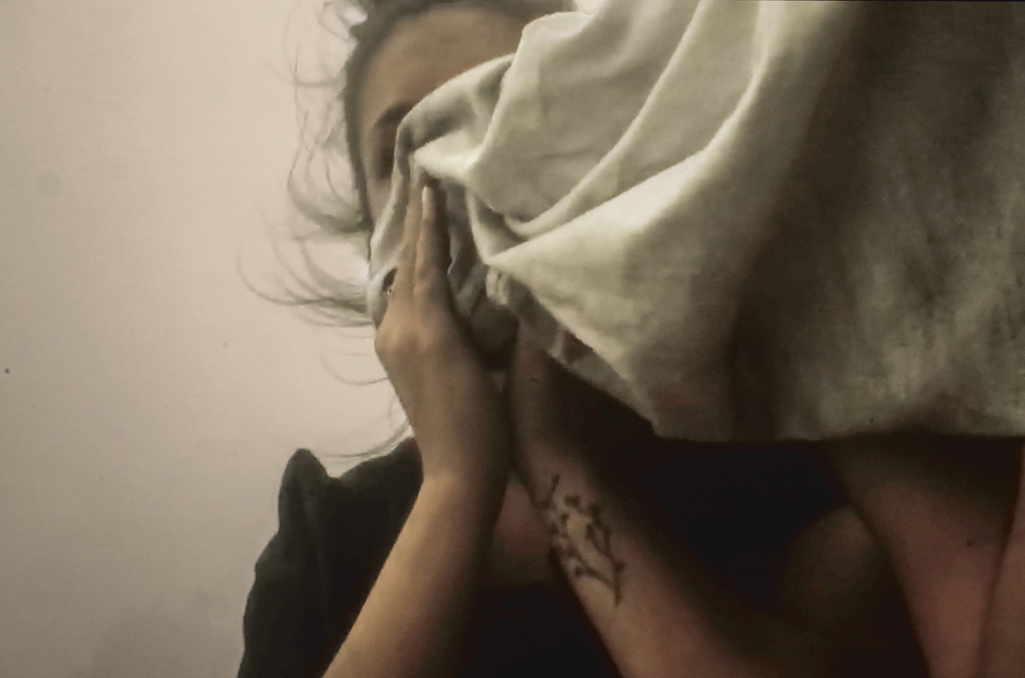 Person with hands covering face, wearing white fabric head covering against pale background with warm flesh tones and dark shadows.