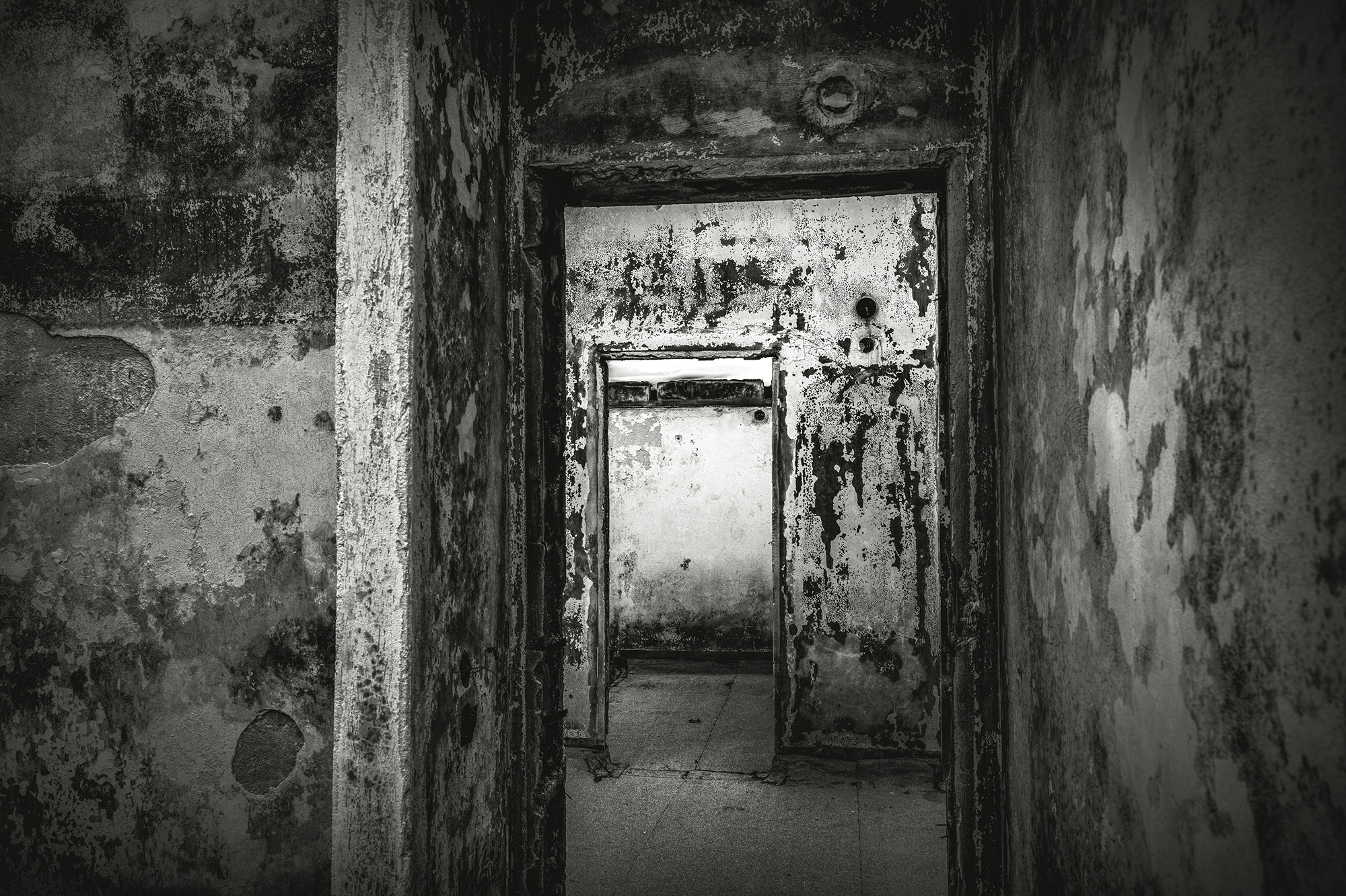 Deteriorated interior room with peeling paint, mouldy walls, and damaged doorframe leading to another dimly lit space.