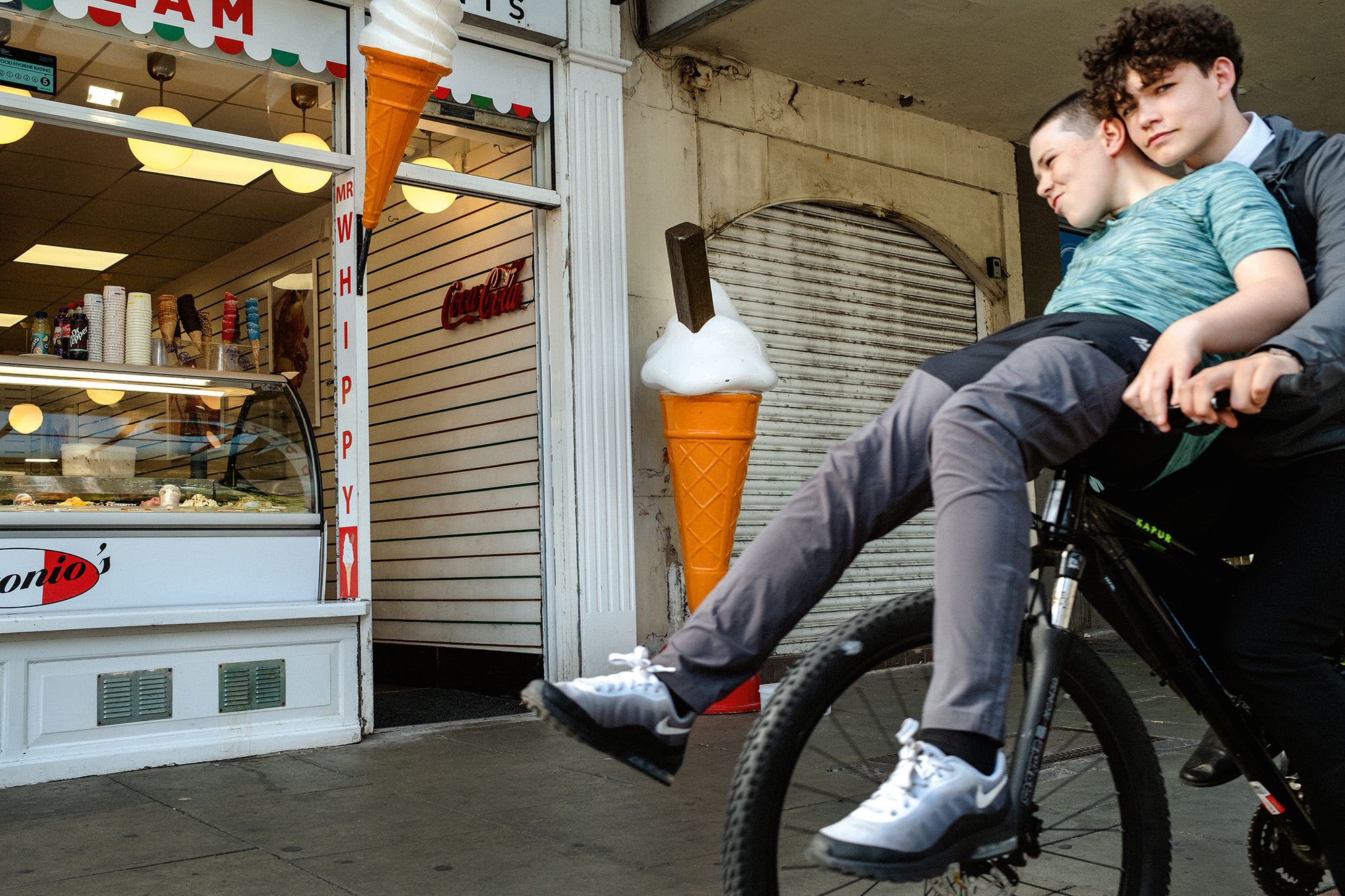 Two teenagers on a bicycle outside closed shops with metal shutters, one wearing a light blue striped shirt, the other in dark clothing.