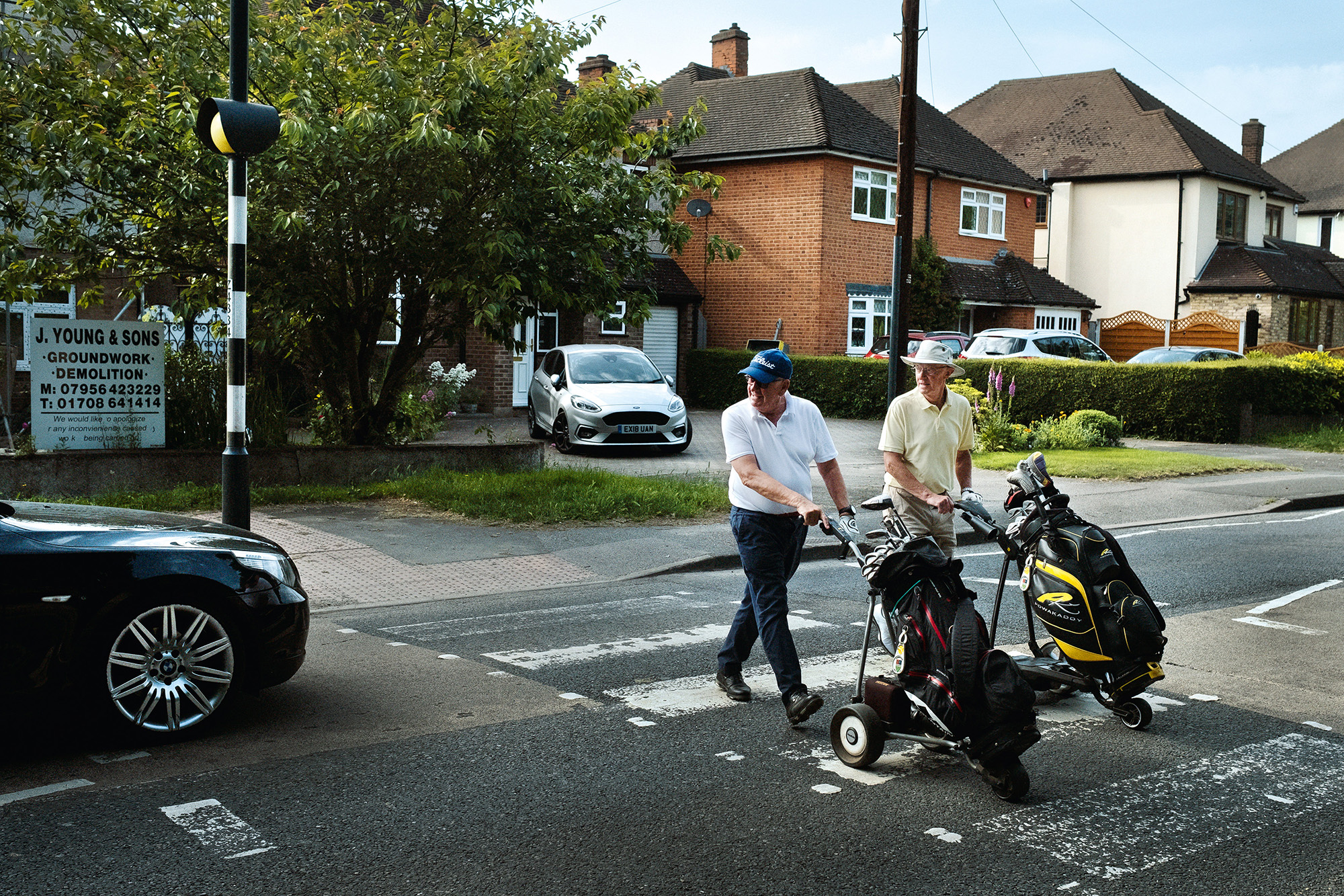 Two golfers with trolleys crossing residential road near brick houses, cars parked, green trees and business sign visible.