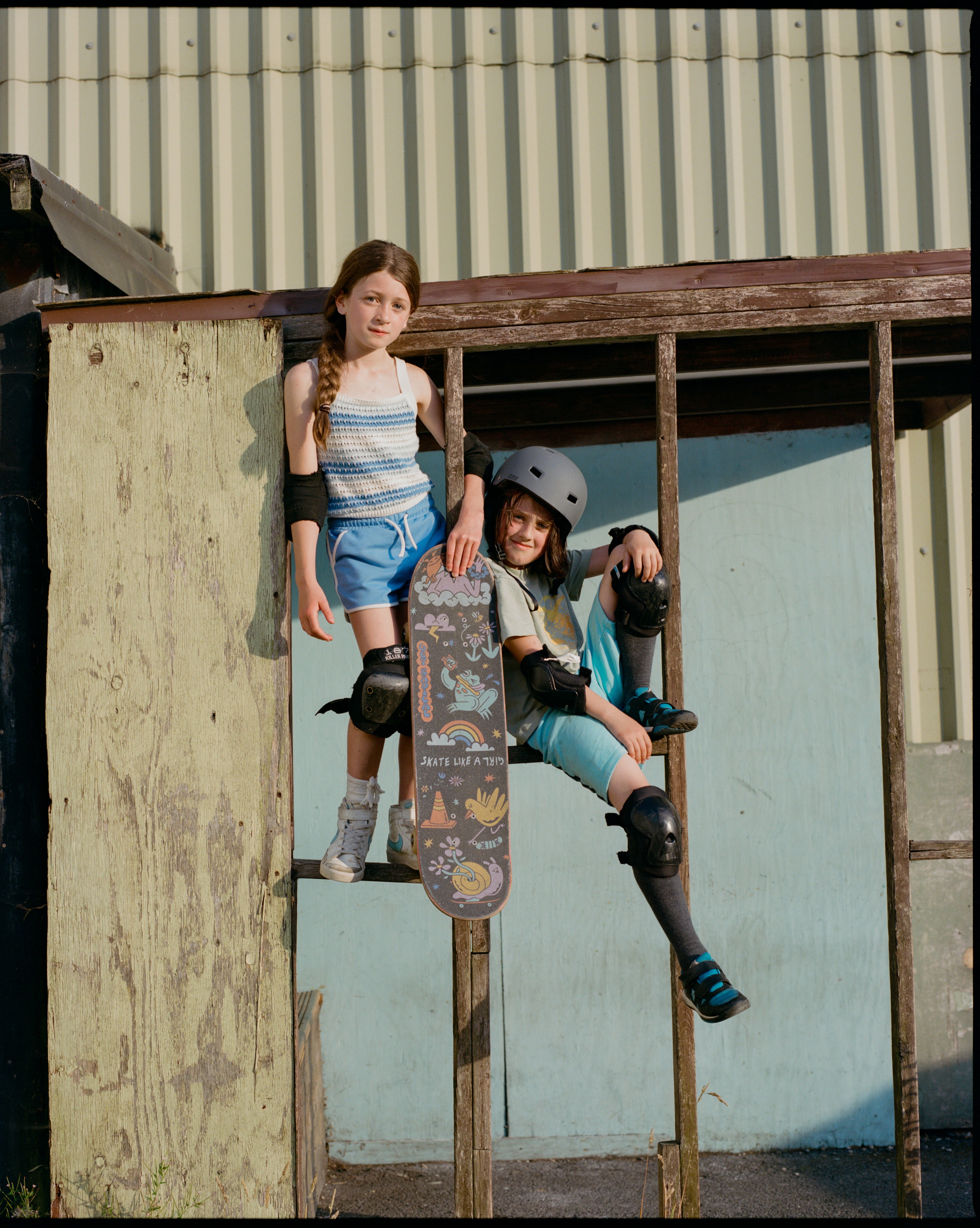 Two young girls sitting on a wooden structure, one holding a skateboard. Colourful clothing, striped and patterned.