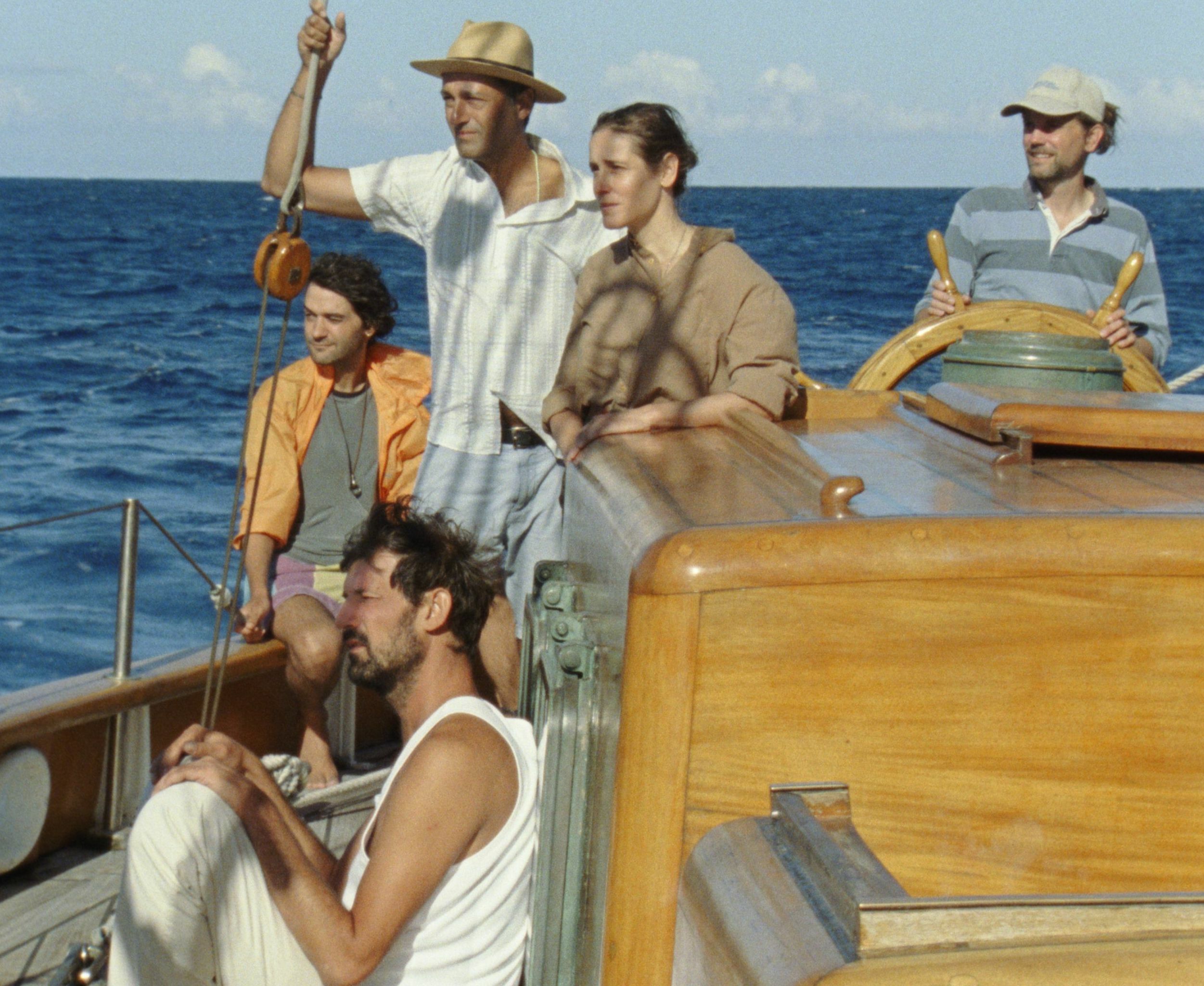 Group of people standing on a wooden boat in the middle of the ocean, wearing casual summer clothing.